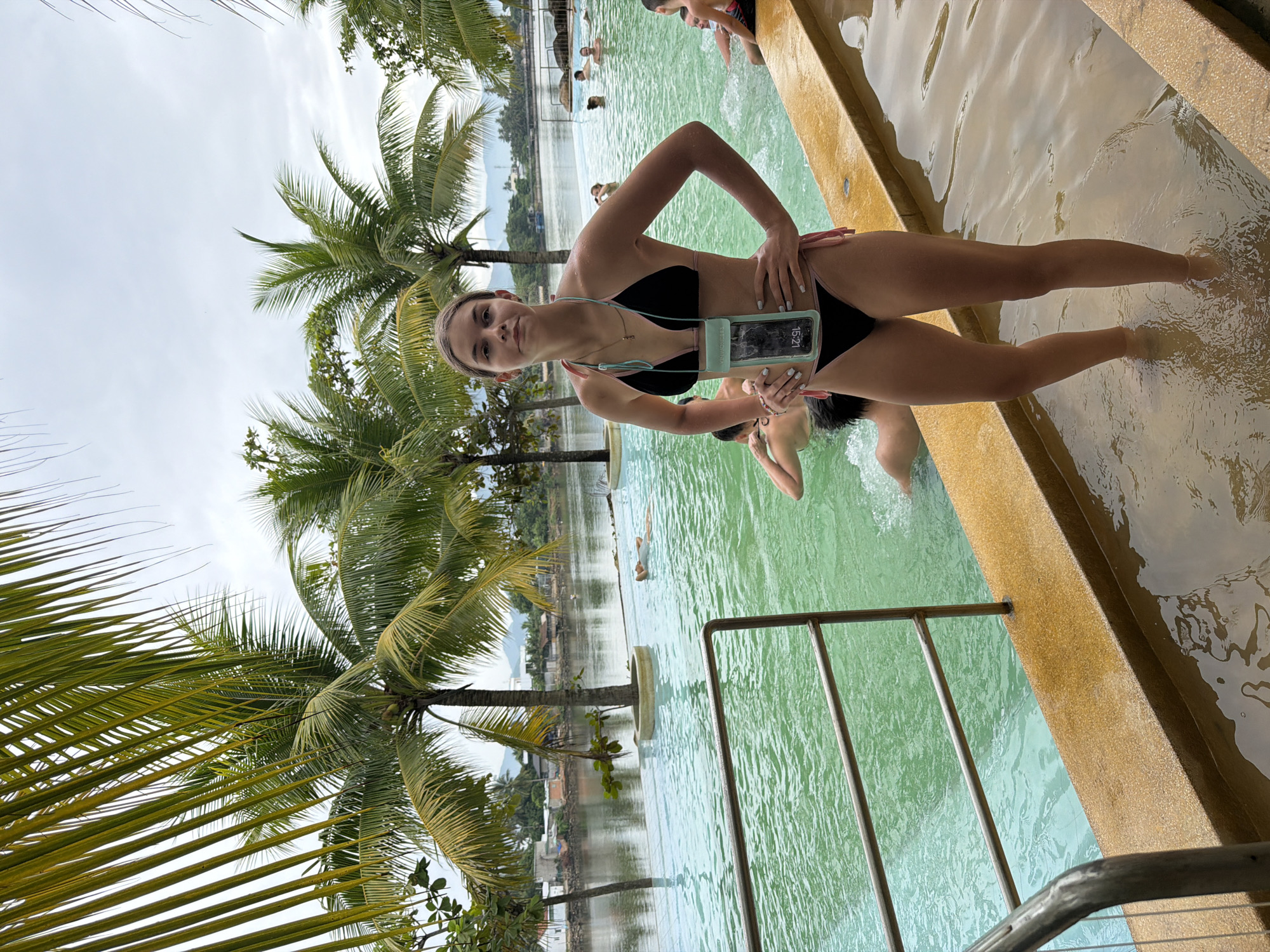 This media file shows a young woman posing for a photo by a pool with lush palm trees and a cloudy sky in the background, suggesting an overcast day. The woman is wearing a black bikini with light pink trim and is holding a waterproof phone case around her neck. She is standing in shallow water next to a textured yellow pool edge. In the background, several other people are visible in the larger pool, some appearing to swim or relax. The overall scene is of a tropical resort or water park. The time on the phone case reads "15:21." The location is identified as Nha Trang, Vietnam, which aligns with the presence of palm trees and a resort-like atmosphere. The woman's expression is neutral, and the image conveys a relaxed, vacation-like mood.