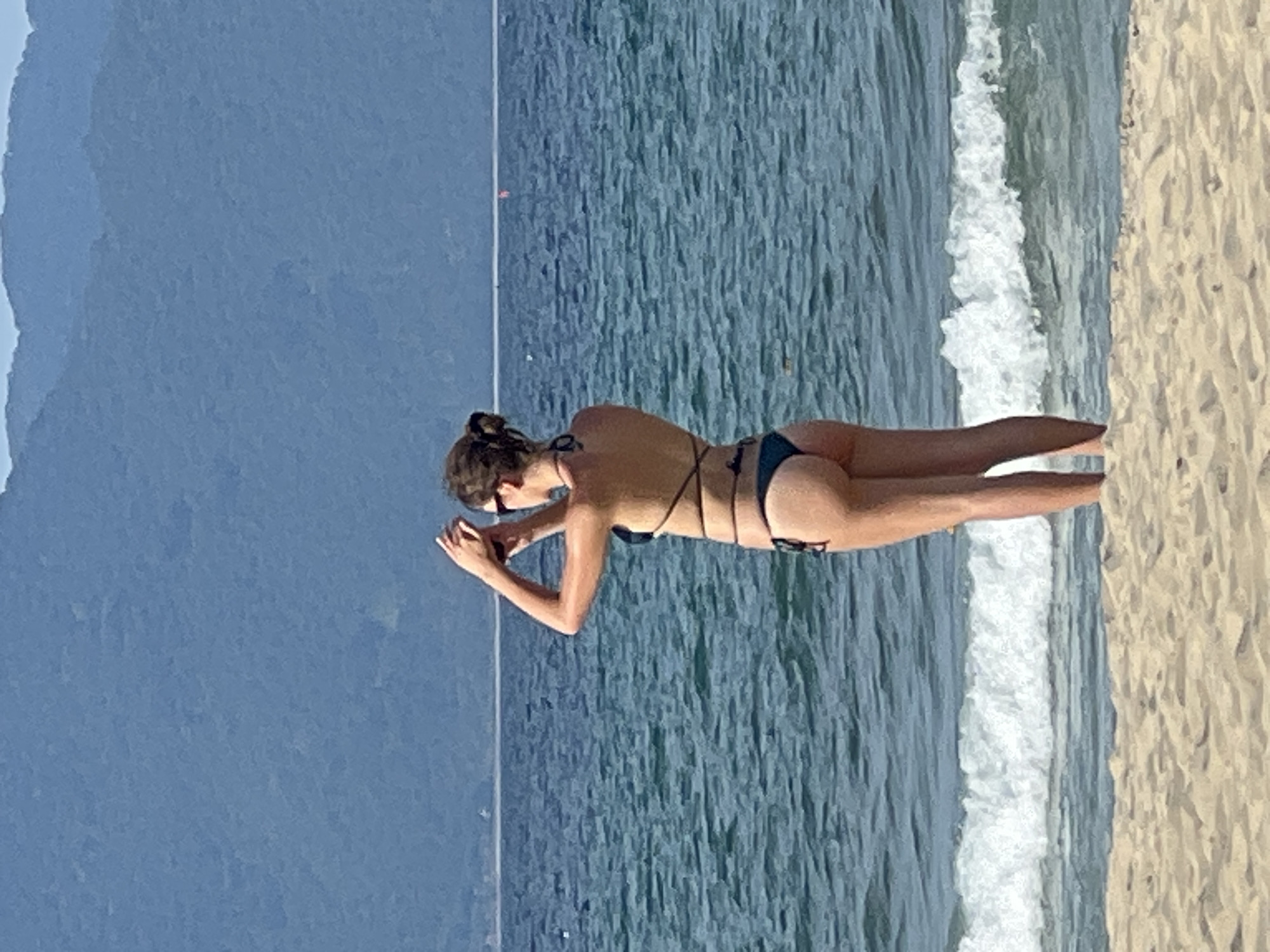 A young woman in a dark bikini stands on a sandy beach, facing away from the camera and towards the ocean. She is holding what appears to be a phone up to her face, as if taking a picture or video. Her hair is pulled back in a bun. The ocean behind her is a deep blue, with small waves breaking on the shore. In the distance, a range of blue-green mountains form the horizon. The scene suggests a sunny day at a coastal location, likely in Đà Nẵng, Vietnam, given the presence of mountains in the background that are characteristic of the region's coastline.
