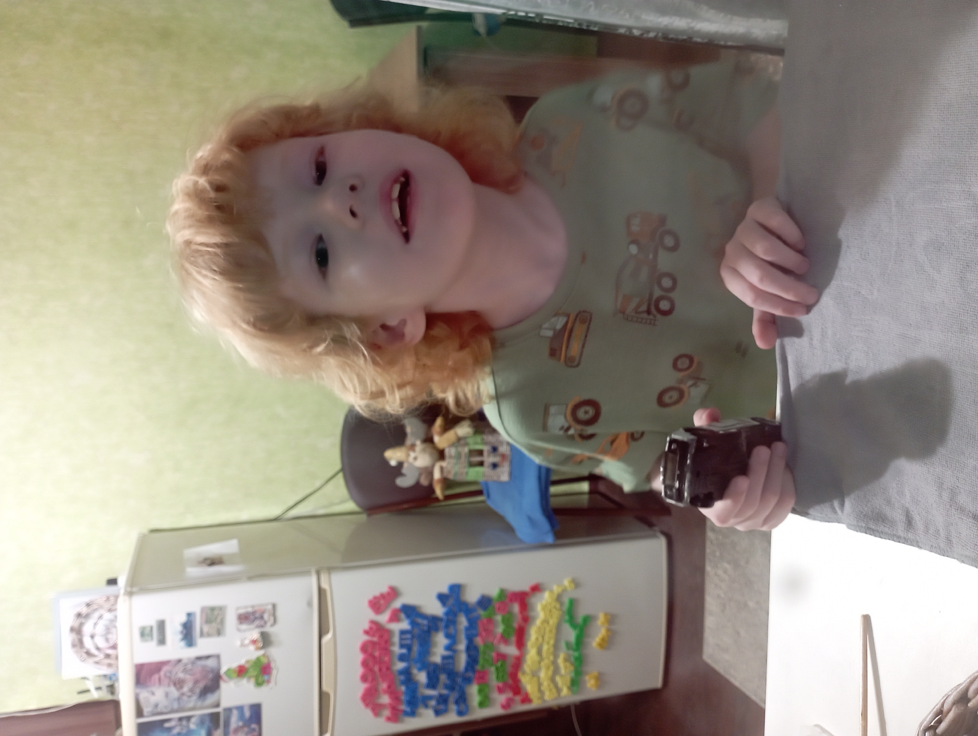 A young child with blonde hair is sitting at a table, looking at the camera. They are wearing a green shirt with images of construction vehicles. The child is holding a small black toy in their right hand. In the background, there is a white refrigerator with colorful magnetic letters and pictures on it, and a stuffed animal on a chair. The room appears to be a domestic setting, possibly a kitchen or dining area. No specific location in Blagoveshchensk, Russia can be determined from the image.