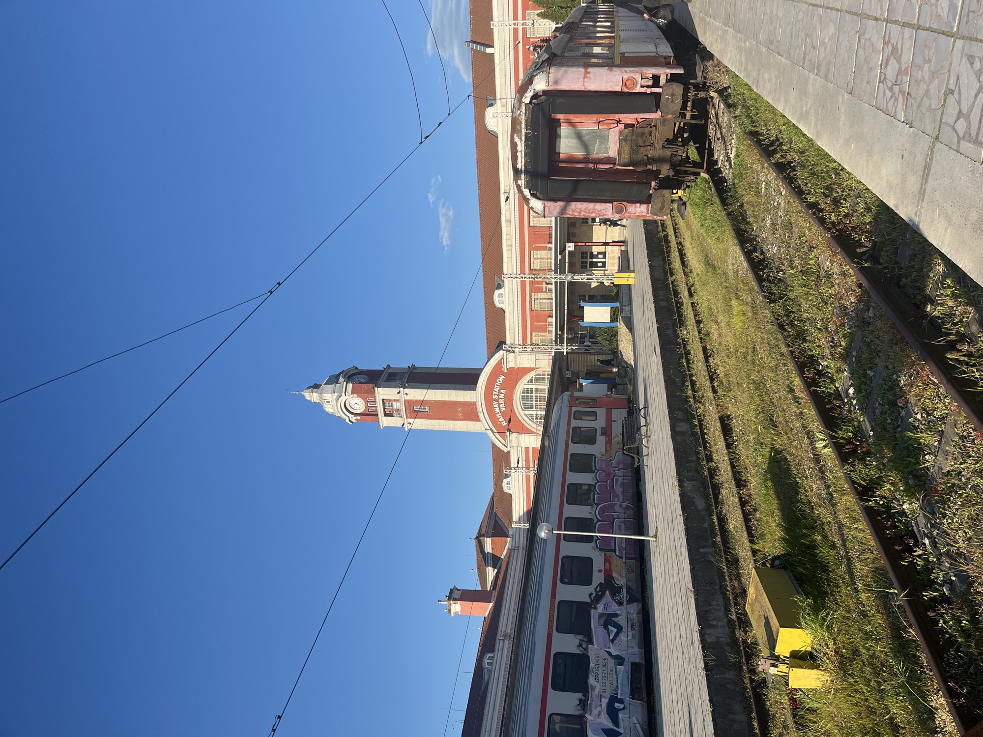 The image depicts the Varna Railway Station in Varna, Bulgaria, under a clear blue sky. The red and white clock tower of the station is prominent. Two trains are visible: one newer train with graffiti and one older, weathered train. Train tracks overgrown with grass run along the side of a paved platform. Overhead power lines are strung across the sky. The station building is partially visible behind the trains. No people are visible.