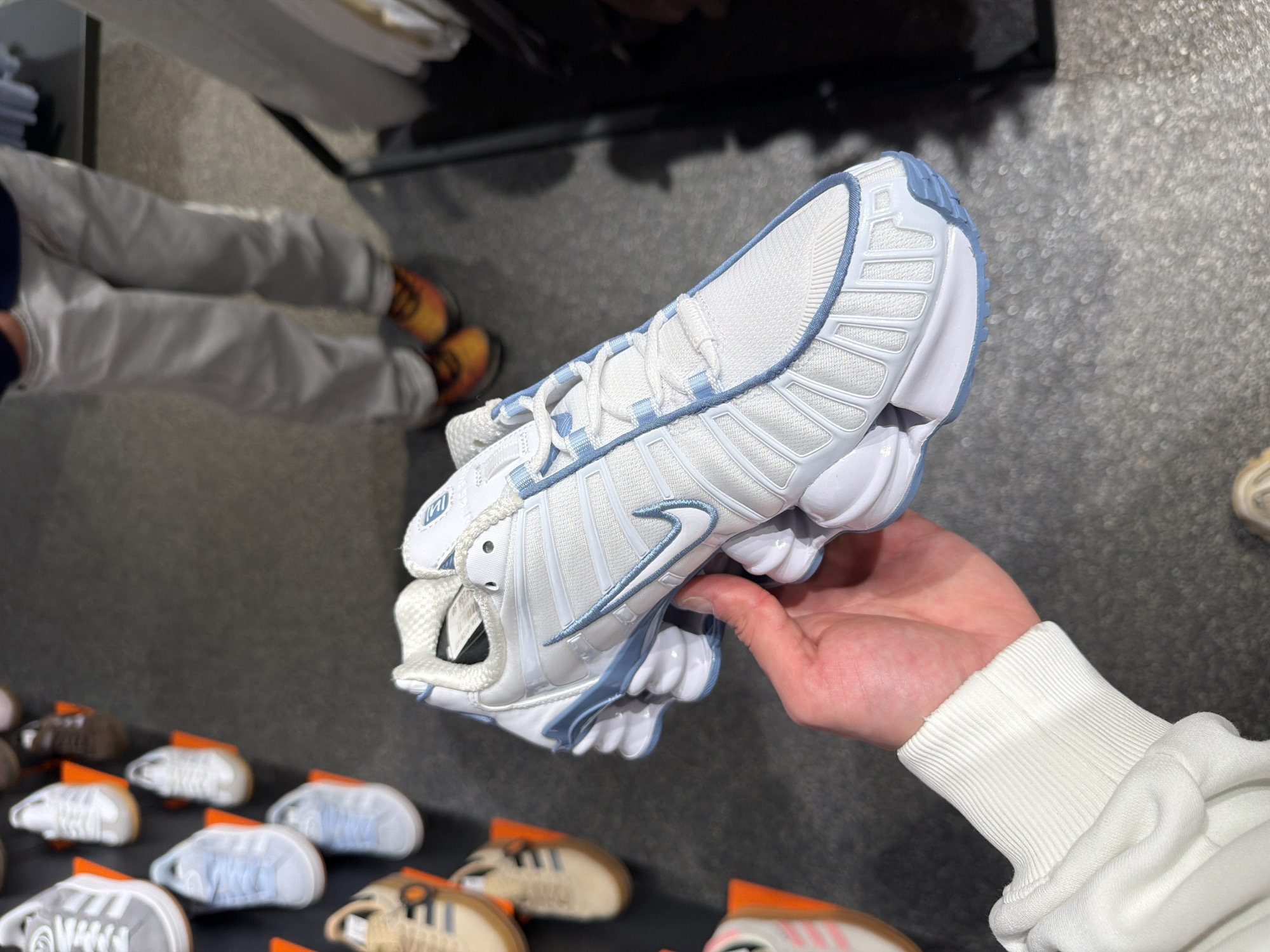 A white and light blue Nike Shox sneaker is held in a person's hand, displayed indoors with other sneakers laid out on a black surface. The setting appears to be a retail store. A person wearing gray pants and orange shoes stands blurred in the background. This image may have been taken in a store in Spreitenbach, Switzerland.