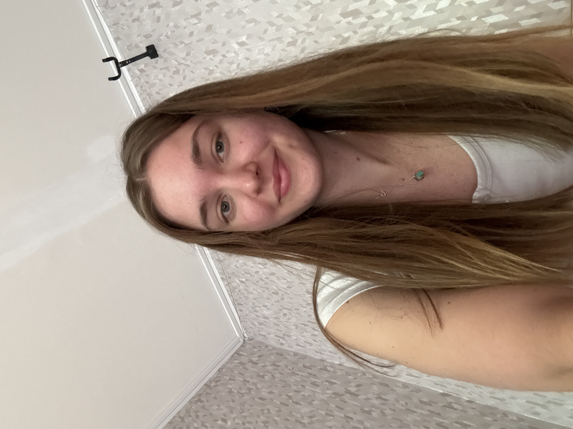 A young woman is captured in a close-up, portrait-style photograph, rotated 90 degrees counter-clockwise. Her long, light brown hair frames her face and shoulders, extending across much of the image. She is looking directly at the camera with a gentle smile, displaying a pleasant and relaxed expression. Her eyes are open, and she wears a simple white top. A delicate silver necklace with a small, round green or turquoise pendant is visible around her neck.
The background indicates an indoor setting, likely a room in Perm, Russia. To the left and top of the frame, there is a plain white ceiling. In the upper left corner, a black, U-shaped hook or bracket is mounted on the ceiling. The right and bottom portions of the image show a wall covered with a light-colored, possibly grey or off-white, wallpaper featuring a subtle, repeating geometric pattern. The lighting is bright and even, characteristic of indoor ambient light. No discernible text is visible in the image.