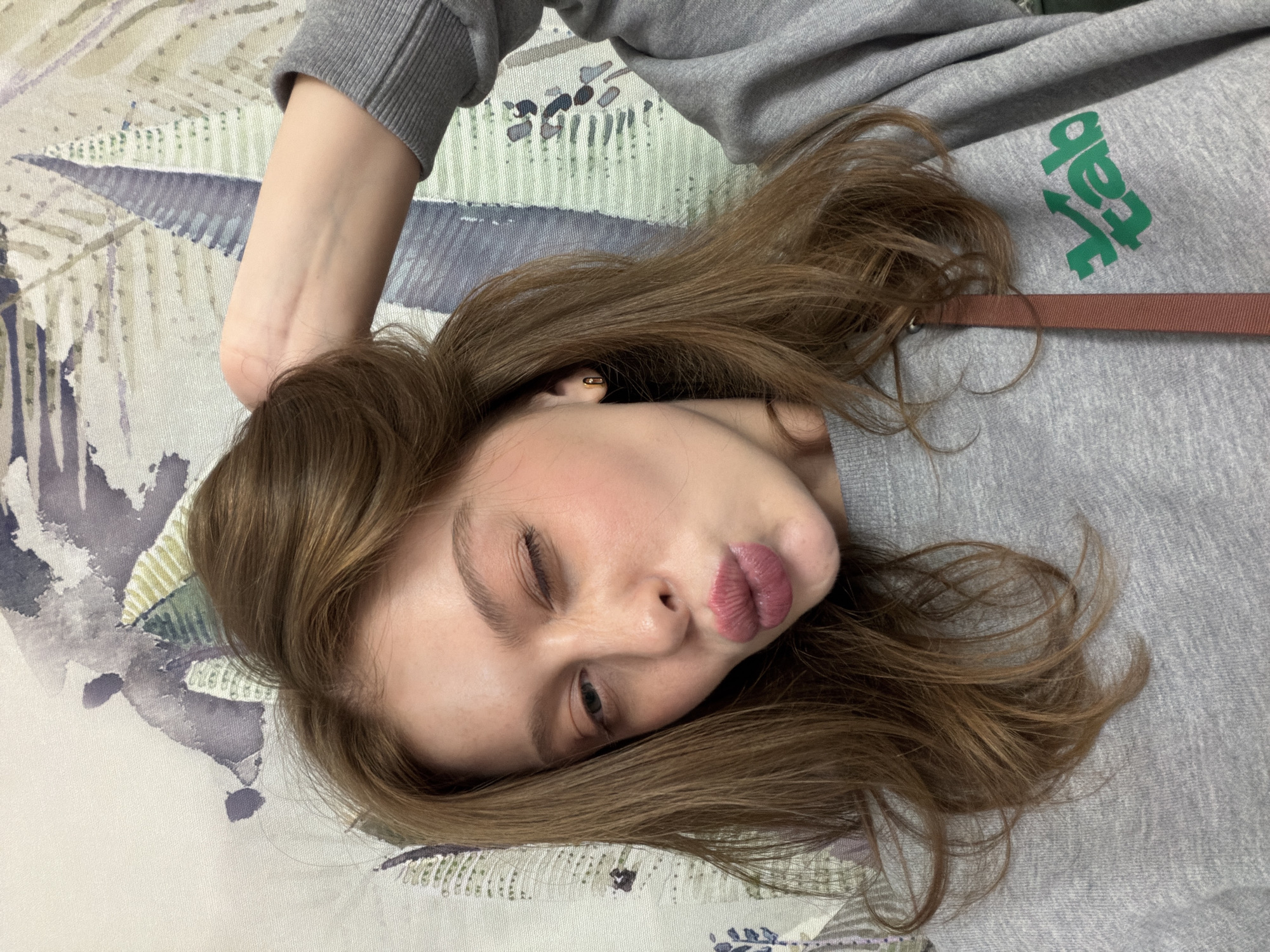 A woman is lying on a floral-patterned surface, likely a bed or similar soft furnishing. She is wearing a grey sweatshirt with green lettering visible, and her brown hair is spread around her head. Her right eye is closed in a wink, and her lips are pursed in a kiss. The background features a light-colored fabric with a watercolor-style floral design in shades of blue, purple, and green. The location is not discernible from the visual information.