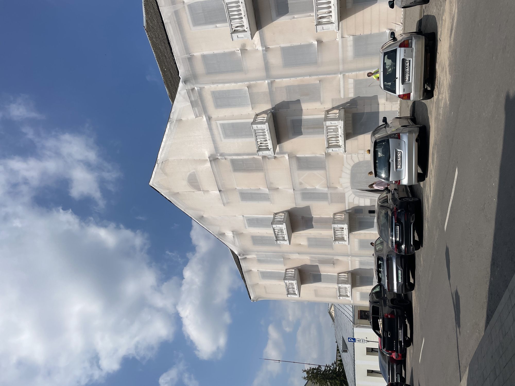 A street view in Hrodna, Belarus, shows a building undergoing renovation, covered in white netting. Several cars are parked along the street in front of the building. The sky is blue with scattered white clouds. The building appears to be a multi-story structure with balconies. License plates are visible on some of the parked cars, indicating a local setting. A few people are walking near the building and cars.