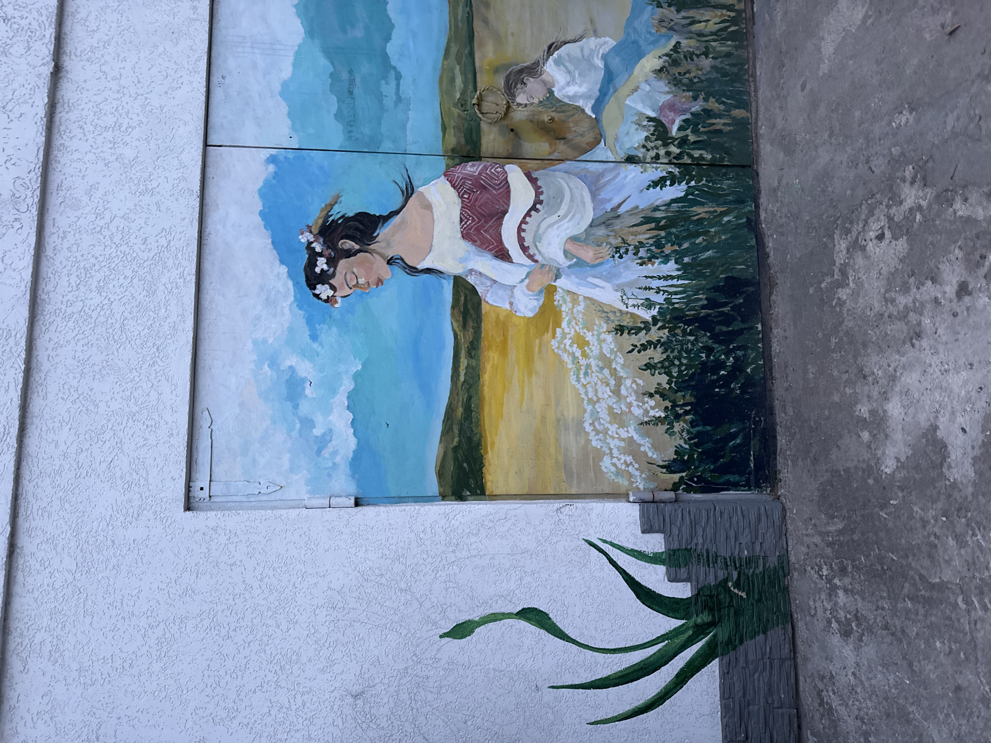 A mural depicting two women in traditional dress, likely in an agricultural or rural setting. The mural is painted on a wall constructed of stucco, with the mural divided into four rectangular panels and oriented vertically. The overall image appears to be a painted mural on an exterior wall, possibly in an urban area, as suggested by the surrounding cement and the construction material of the wall. Details of city location are not discernible.
