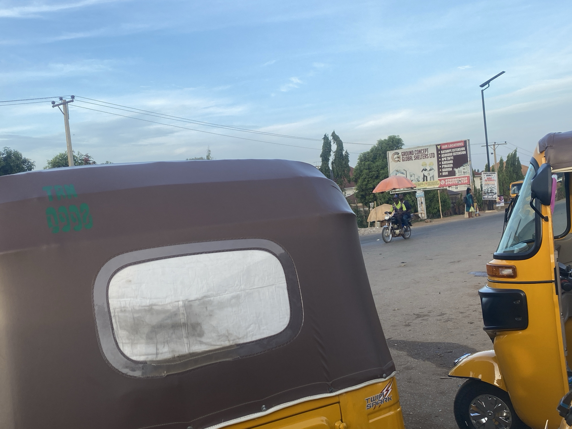 The image depicts a street scene in Galadimawa Village, Nigeria, with two yellow auto rickshaws (tuk-tuks) in the foreground. The left tuk-tuk is partially visible, showing its brown canvas roof with a small rectangular window and the text "TWIN SPARK". The tuk-tuk on the right is fully visible, including its windshield and part of the wheel.
In the background, a motorcyclist with a passenger is riding on the road. A large billboard for "GROUND CONCEPT GLOBAL SHELTERS LTD" is visible, listing several locations including Katampe Extension, Asokoro Extension, Apo Tafi, Karsana, Pyakasa, Kabusa, and Kuje, along with phone numbers. Another sign for "COPA CABANA HOMES" is partially visible as well. A person is walking on the side of the road. Utility poles and overhead power lines are also present. The sky is a light blue with scattered clouds.