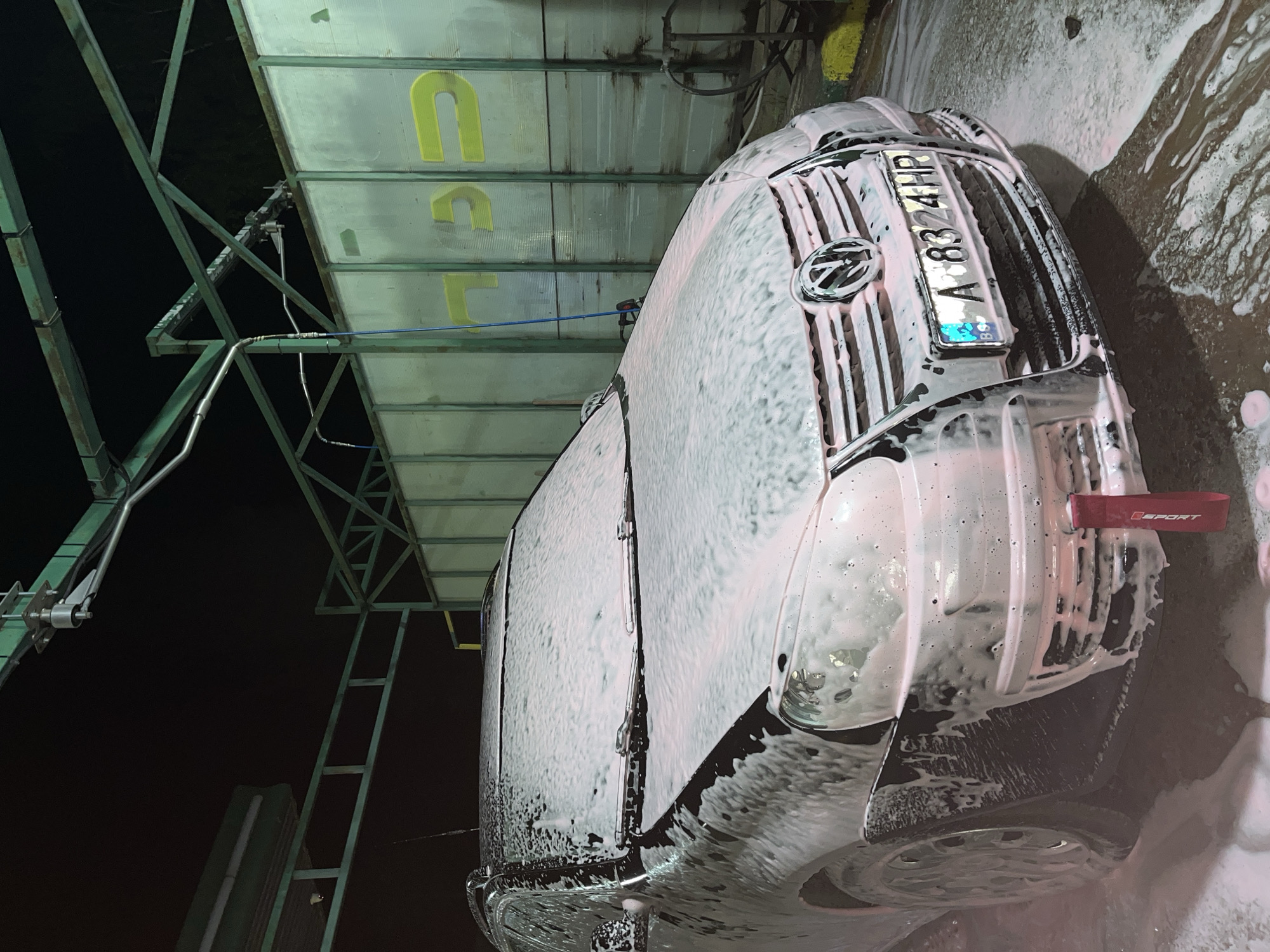 A close-up, low-angle shot captures a dark-colored vehicle, likely a van, undergoing a car wash. The vehicle is heavily coated in white foam, which is tinged with pink in some areas. Prominently displayed on the front grill is a license plate reading "A 8324 HP," and a Volkswagen logo is visible. The environment suggests a car wash facility, with metal framework visible above and to the left. Yellow lettering is seen on a structure behind the vehicle. The image is taken at night, evident by the dark sky. The presence of a detail reading "SPORT" suggests that the vehicle may have some enhanced features. The location is Sungurlare, Bulgaria.