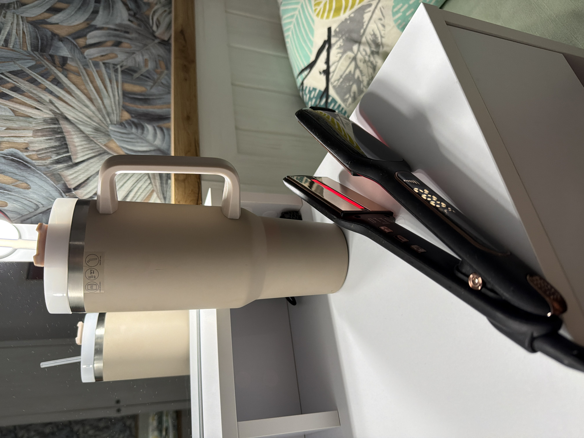 This image depicts an indoor scene located in Plovdiv, Bulgaria. The primary subjects are a large beverage tumbler and a hair straightener, positioned on a white, flat-surfaced piece of furniture, likely a vanity or bedside table.
The tumbler, light beige in color, is made of what appears to be stainless steel, featuring a prominent handle on its right side and a white lid. A small, light-brown object protrudes from the lid, and a reflection in the mirror behind shows a straw within the tumbler. Three circular icons are visible on the tumbler's surface: one depicting a wash symbol, one indicating "70°C HOT 7H COLD 10H," and another showing a stylized cup.
Behind the tumbler, a rectangular mirror reflects its back and a portion of the room. The background wall is covered with wallpaper showcasing a pattern of large, stylized tropical leaves in shades of grey, blue-green, and brown. A wooden ledge runs horizontally across the wall beneath the wallpaper. Further to the right, a white paneled structure is visible, potentially a headboard or wainscoting.
In the foreground, a black hair straightener with rose gold or copper accents rests open on the white surface. Its heating plates are illuminated with a distinct red glow, indicating it is powered on and actively heating. Control buttons and a display panel are visible on the top plate. The power cord extends out of frame to the left.
To the far right, a pillow with a light blue-green and yellow leaf pattern is partially visible, suggesting a bed is situated nearby. The white furniture piece supporting the tumbler has an open shelf or cubby underneath. No individuals are present in the scene. The overall lighting is bright, casting subtle shadows on the white surface.
