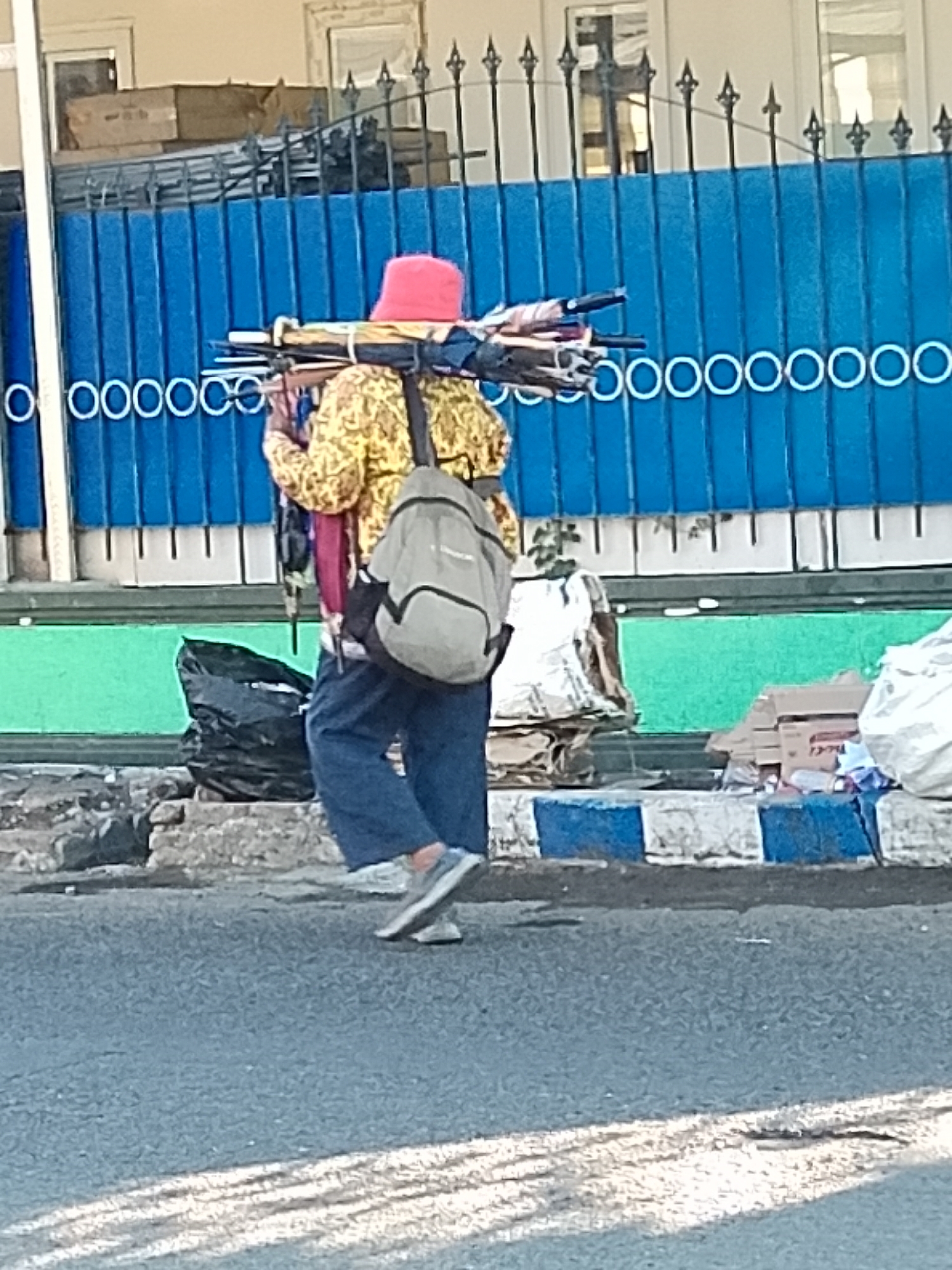 A person with a red hat and a yellow patterned shirt walks on a street in Surabaya, Indonesia, carrying a bundle of umbrellas on their shoulders, and a backpack. The person is wearing blue pants and gray shoes. The background features a blue metal fence, a green wall, and some debris on the side of the road. The lighting suggests it is daytime.