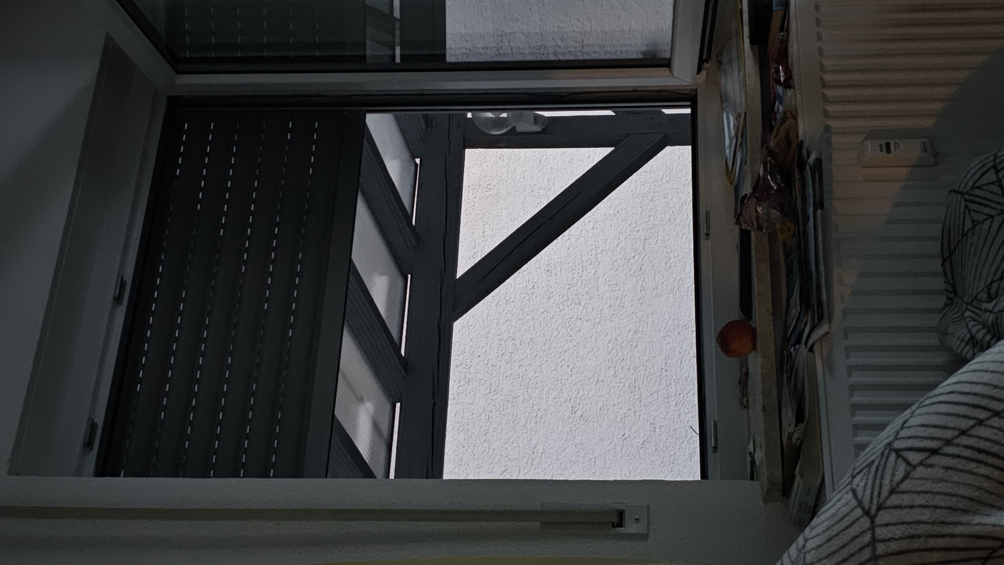 A dark, partially open window reveals a rough-textured white wall outside, partially obscured by a grey, angled structure with a translucent, ribbed roof. The bottom of the window displays a windowsill cluttered with various objects including papers, what appears to be a peach, and a crumpled wrapper. Below the windowsill is a white radiator, and in the foreground, a pillow with a black, geometric pattern is visible. The scene suggests an indoor setting looking outwards.