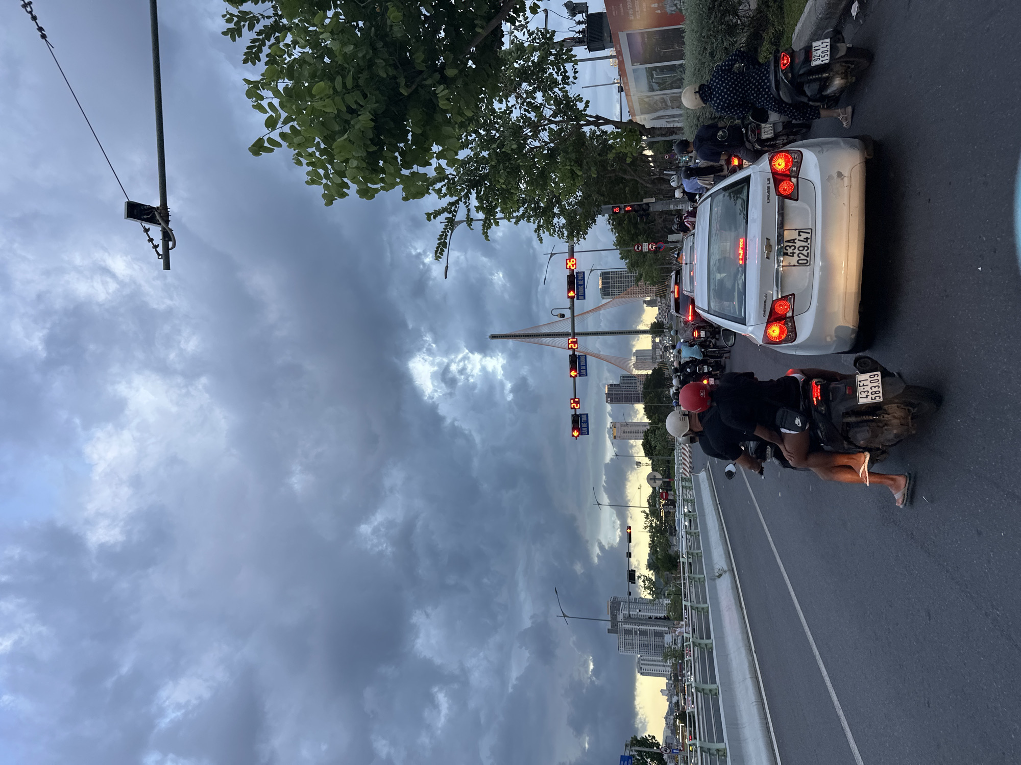 The image depicts a traffic jam on a city street in Đà Nẵng, Vietnam, under a cloudy sky with dramatic cloud formations. In the foreground, several motorcycles and a white Chevrolet car are stopped in traffic. A couple on a motorcycle in the middle distance is visible from behind, with the passenger wearing a red helmet and the driver a black helmet. The car in front has a license plate that reads "43A 02947." Further down the road, more vehicles and people are present. In the background, tall buildings and a distinctive cable-stayed bridge are visible, illuminated by the dim light of dusk. Traffic lights are also visible, some displaying red. Lush green trees line the right side of the road. The overall atmosphere suggests a busy urban environment during a transition in weather conditions.
