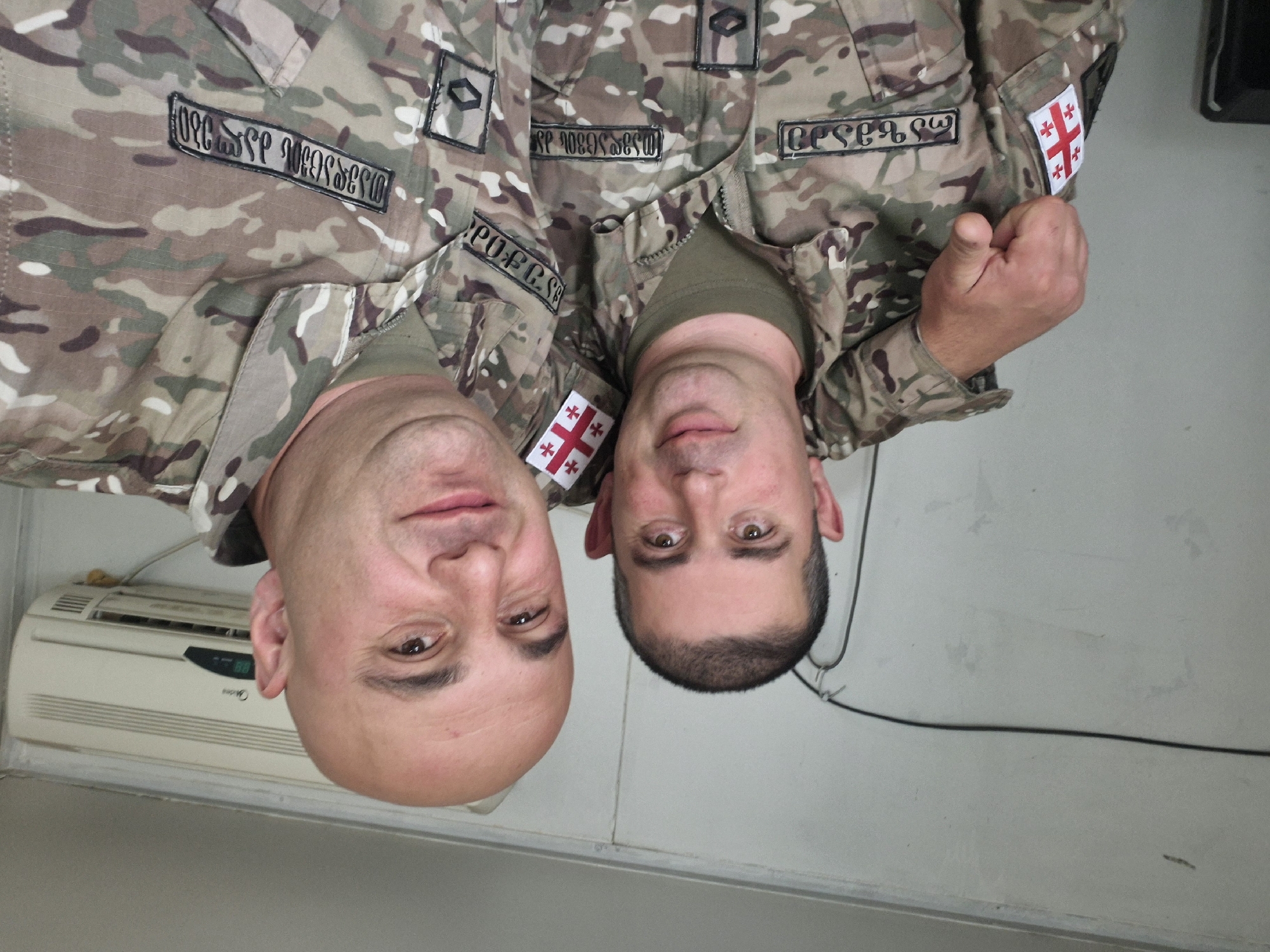 Two men in military camouflage uniforms with Georgian flag patches are standing together indoors. The man on the left is bald, and the man on the right has short dark hair. The uniforms have name tags in Georgian script. The background appears to be an interior room with a wall-mounted air conditioning unit. The image likely depicts the subjects in Tbilisi, Georgia, based on the flag patch and the language on the uniforms.