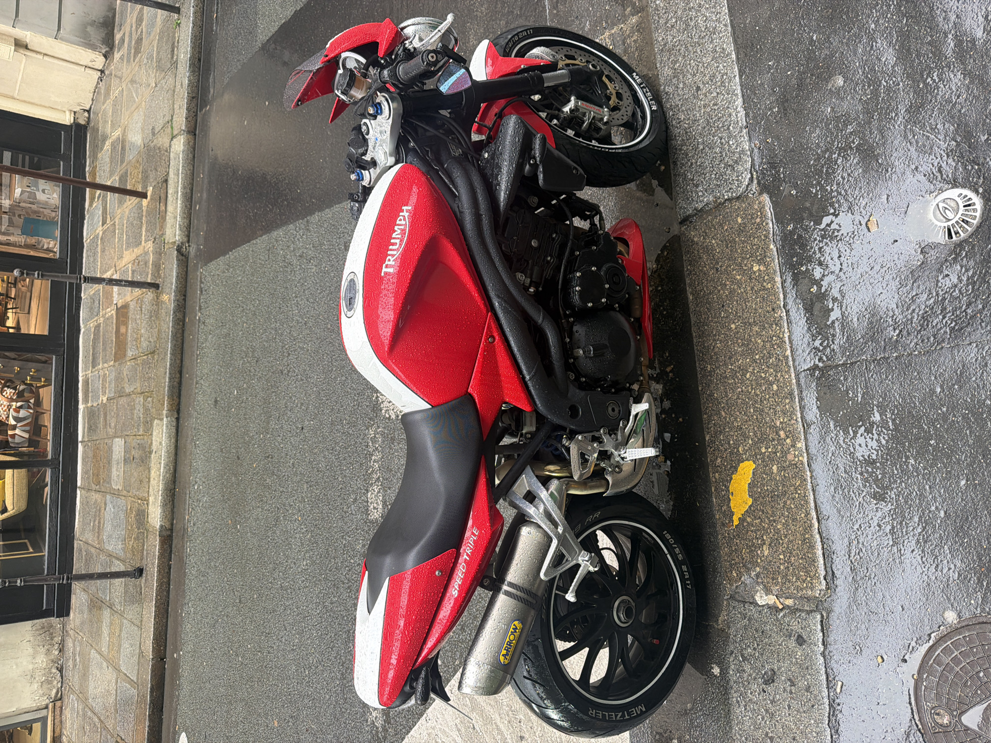 A red and white Triumph Speed Triple motorcycle is parked on a wet street. The street is likely in Paris, France, as evidenced by the architecture of the building to the left of the image. The motorcycle is parked next to a curb, and there is a manhole cover on the street nearby. The motorcycle appears to be wet, likely due to recent rain. A store front is visible next to the street on the left.