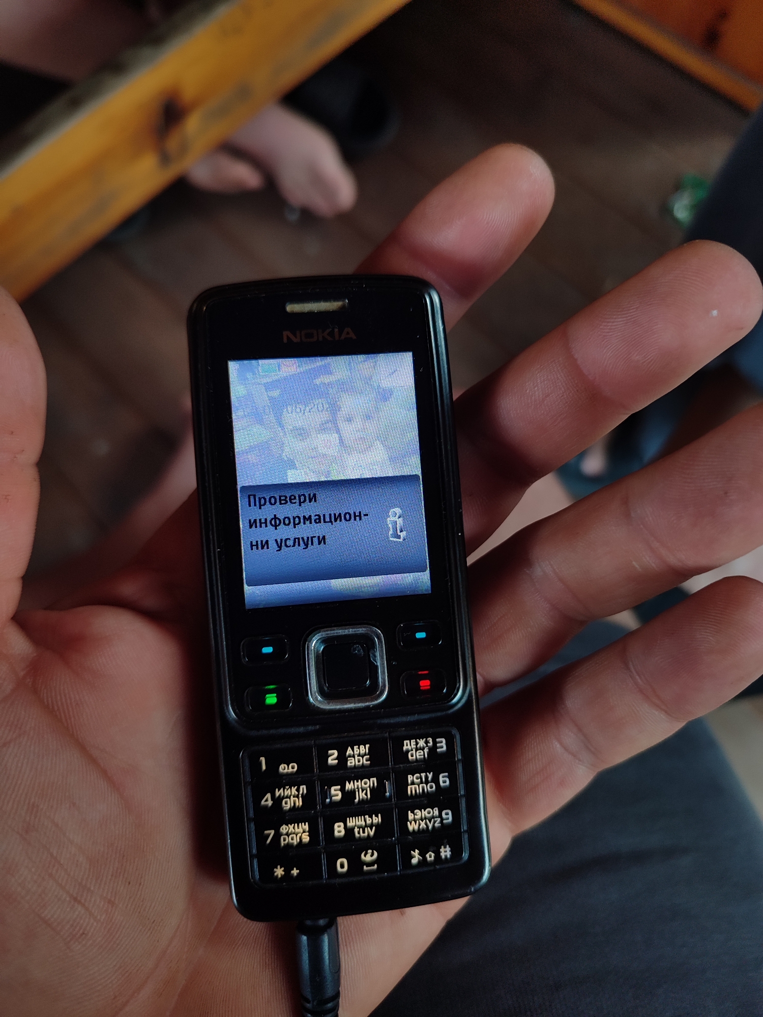 A close-up, high-angle shot depicts a left hand holding a black Nokia feature phone. The phone is active, displaying "NOKIA" branding above an illuminated screen. The screen shows a blue-tinted background image, partially obscured by a rectangular dark blue-grey text box. Within the text box, Cyrillic text reads "Провери информационни услуги" (Verify information services), accompanied by a small white icon on the right, resembling a stylized person or service symbol with an upward arrow. The keypad below the screen features alphanumeric keys (1-9, *, 0, #) with both Latin and Cyrillic script. Green call and red end-call buttons are illuminated. A central silver navigation pad is present. A black charging or data cable is connected to the phone's bottom port. The hand holding the phone has visible skin texture and fingerprints. The background is significantly out of focus, revealing a light brown wooden floor and parts of a blurry yellow-orange wooden structure in the upper left. In the lower background, two bare feet or feet in light footwear are visible. The scene is located in the city of Varshilo, Bulgaria.