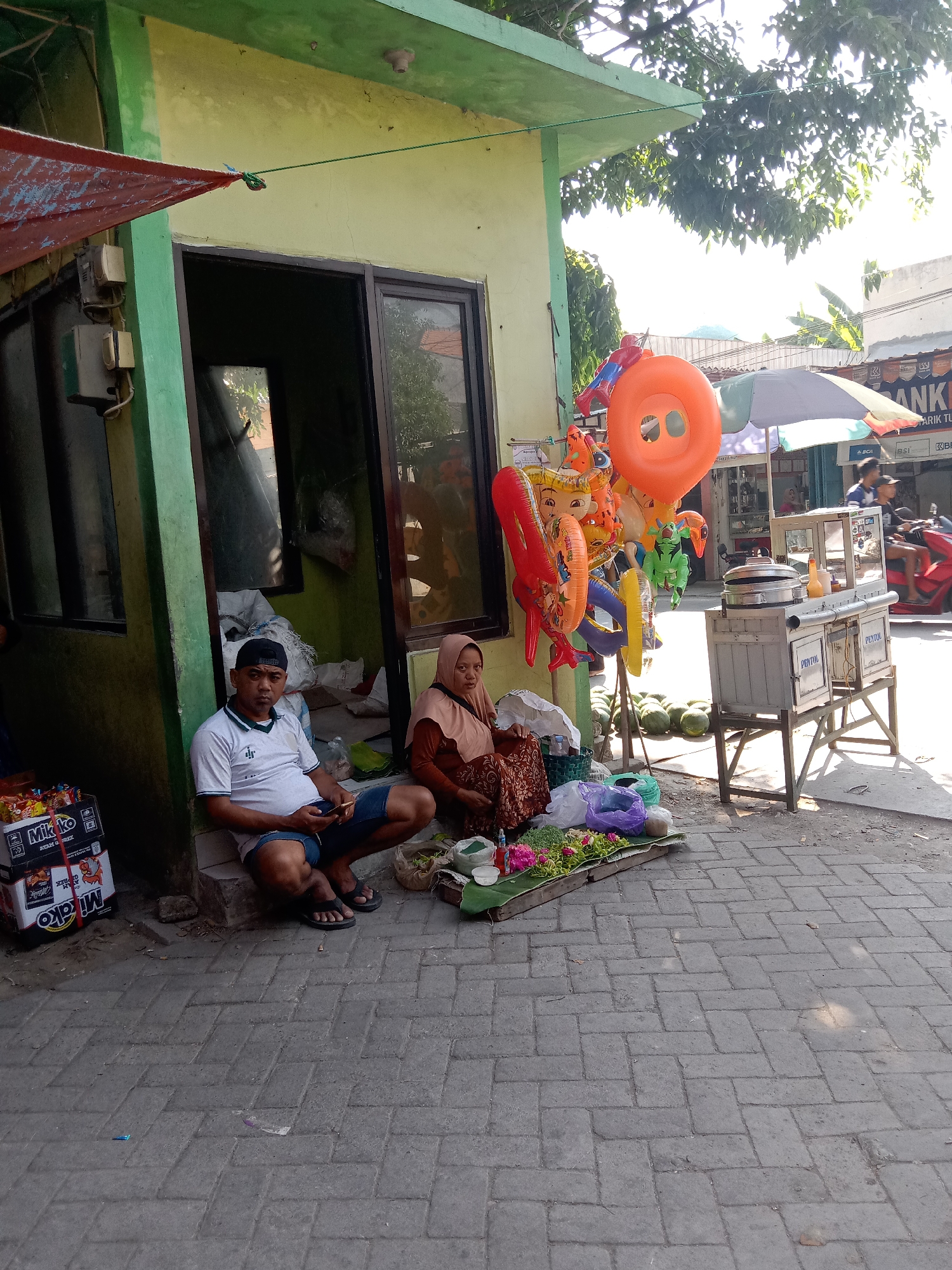 In Sugihwaras, Indonesia, on a sunny day, a man and a woman are seated on a paved walkway outside a small building. The man, wearing a white polo shirt and blue shorts, is looking down at his phone. The woman, dressed in a brown hijab and a patterned skirt, is looking towards the viewer. They are positioned near a display of fresh produce and colorful inflatable toys. A food cart, a motorcycle, and a building with a "BANK" sign are visible in the background. The scene appears to be a street-side market or food stall area.