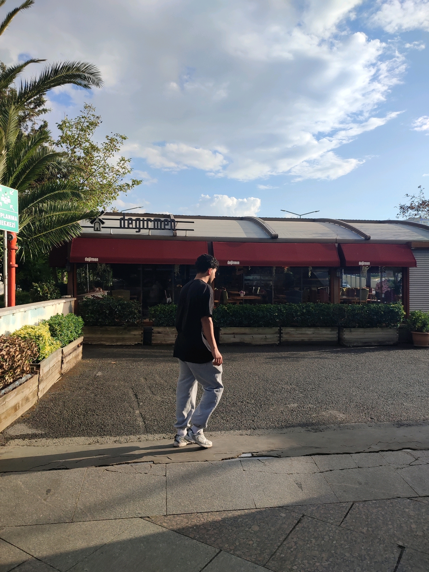 A young person with dark hair is walking from left to right in the foreground, casting a long shadow on the pavement. They are wearing a black t-shirt, grey sweatpants, and white athletic shoes. Behind them is a building with a red awning that reads "Değirmen" in white text. The building appears to be a restaurant or cafe. In front of the building are planters filled with green bushes and some flowering plants. To the left of the building, a palm tree is visible, along with a green sign with white text and a pictogram of people. The sky is blue with scattered white clouds. The ground consists of asphalt and pavement. The scene appears to be outdoors in an urban setting, possibly in Kadıköy, Turkey, given the presence of "Değirmen," a known establishment in that area.