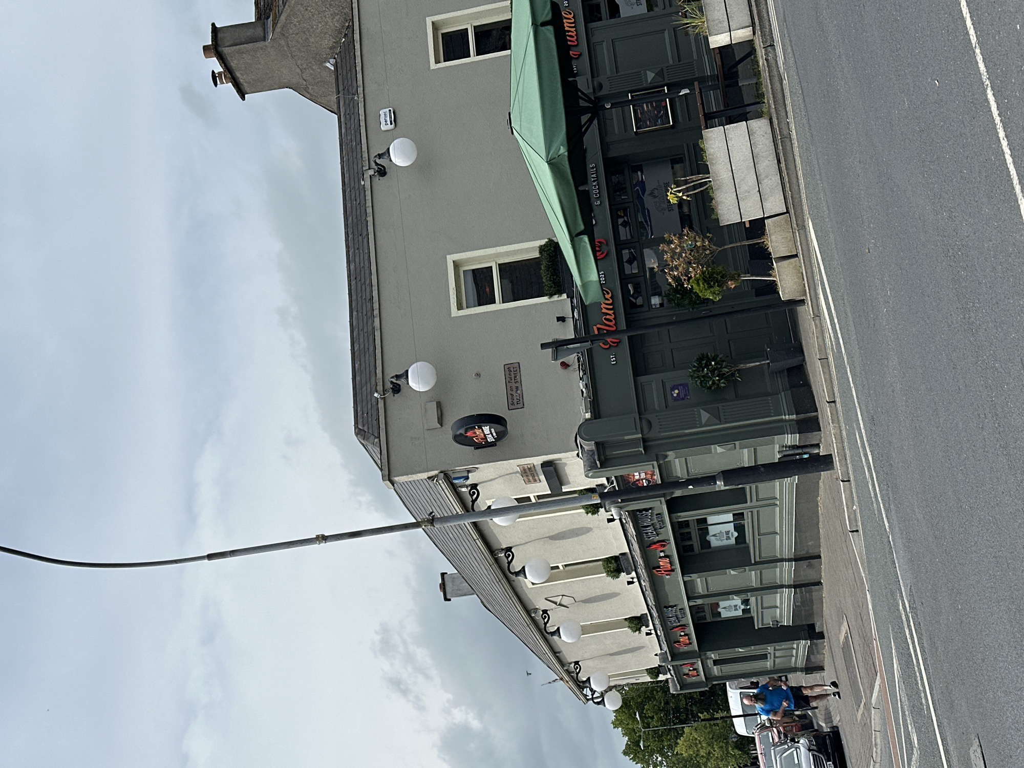 The image depicts the exterior of "Flame Steak House" located in the city of Carlow, Ireland. The building is a two-story structure with a light grey facade on the upper level and a dark green lower level that houses the restaurant. Several round, white outdoor lights are mounted on the upper facade. A green umbrella shades an outdoor seating area in front of the restaurant.
On the sidewalk in front of the restaurant, a man with a beard and blue t-shirt is jogging towards the right side of the frame. He is wearing dark shorts and athletic shoes. Several cars are parked or driving on the street in the background. The sky is overcast with grey clouds. A lamppost is prominently positioned in the foreground, leaning slightly to the left. A chimney with a brown cap is visible on the roof of the building. The street in front of the building appears to be a public road with white lane markings. The overall impression is a daytime street scene in an urban environment.