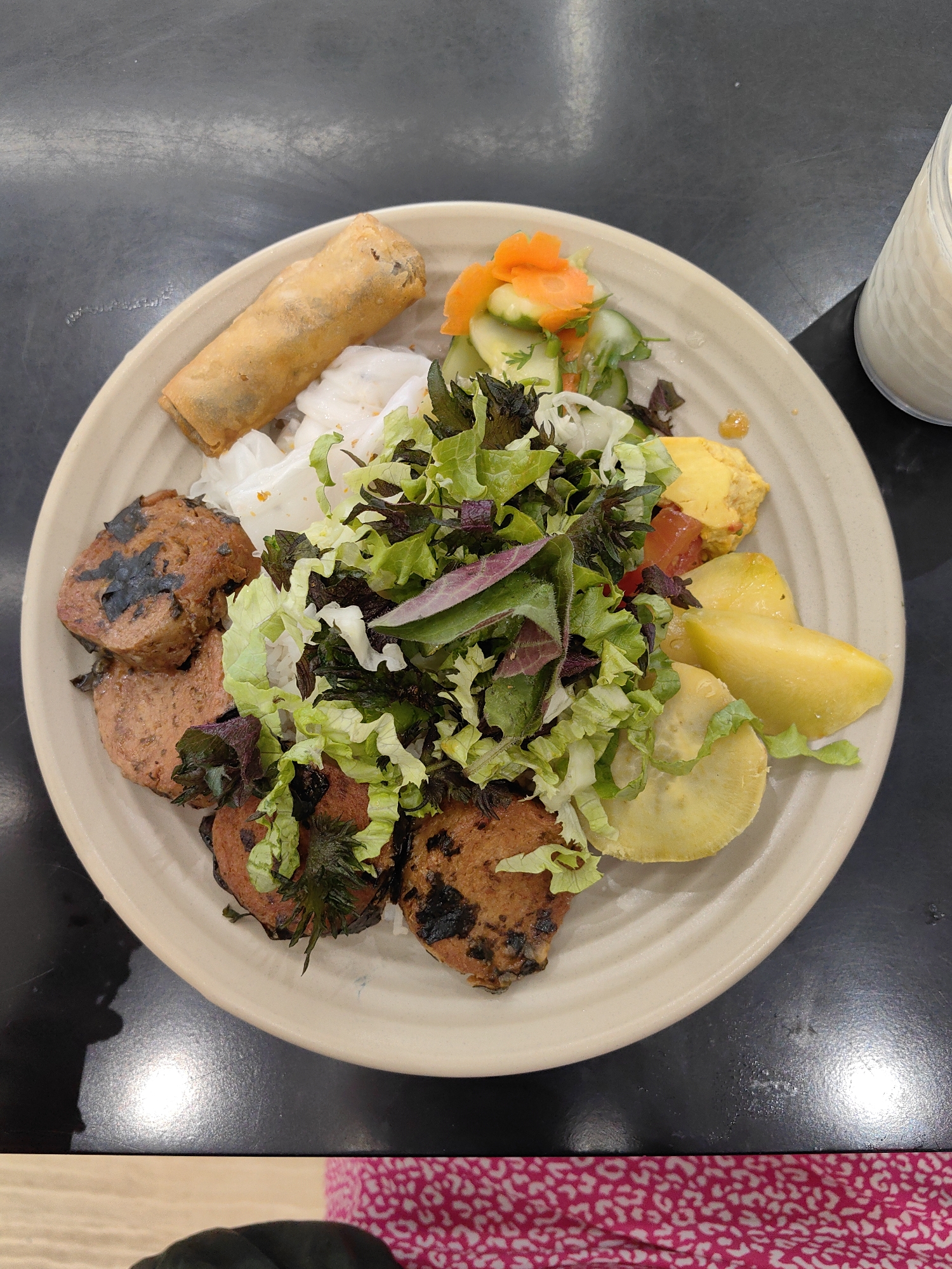A top-down, close-up shot presents a plate of food. The plate is beige with a subtle concentric circle design. The food includes a spring roll, sliced cucumbers and carrots, lettuce, what appears to be rice noodles, cutlets with what looks like burned bits on them, and pieces of what seems to be yellow tomato and some yellow vegetables. A white beverage in a glass is positioned near the upper right side of the plate. The plate sits on a black tabletop. The bottom of the frame reveals a person wearing black gloves and a patterned shirt in pink and white. This image was likely taken in Hong Gai Ward, Vietnam.