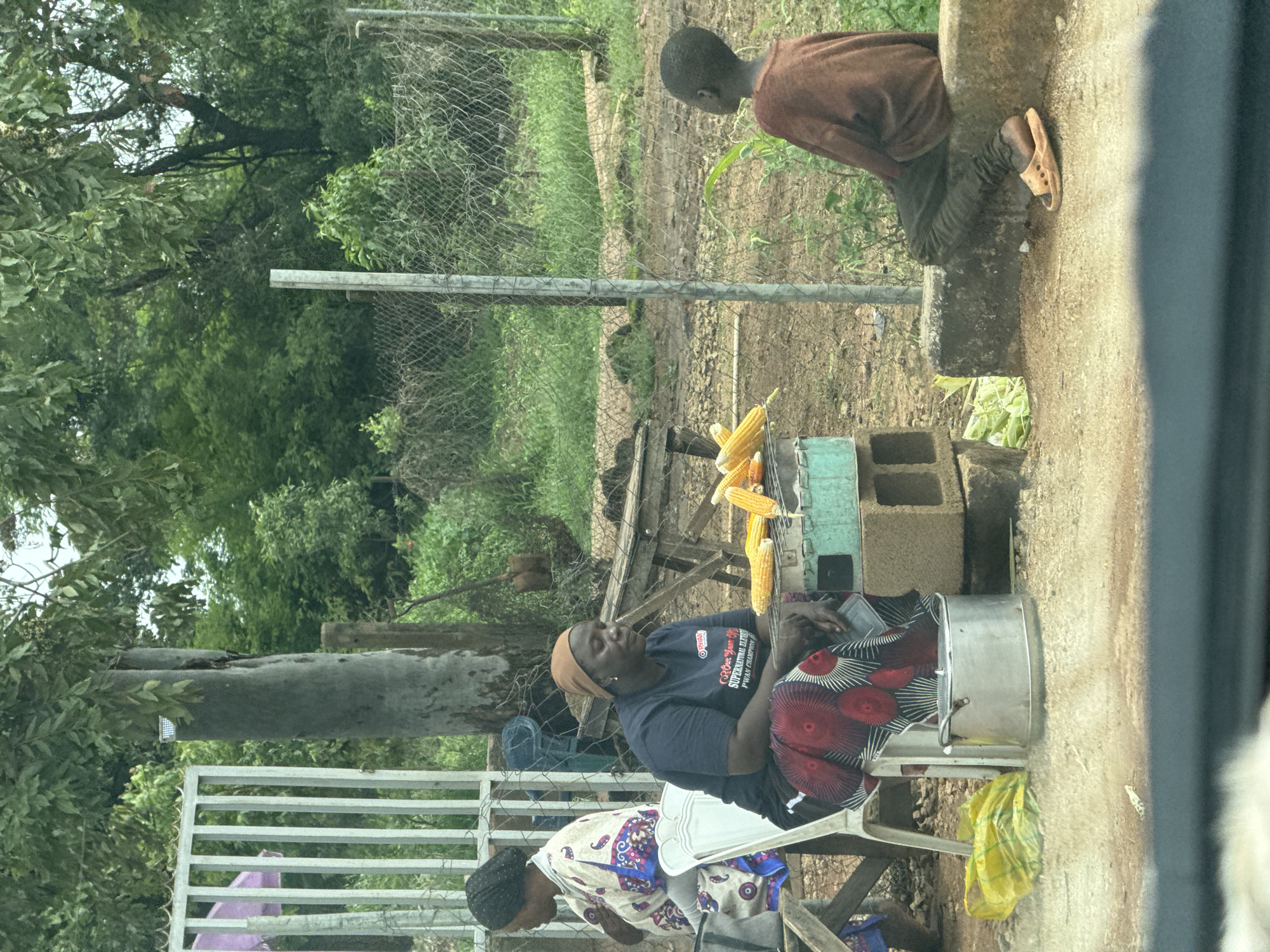 The image depicts an outdoor scene in Gura Top, Nigeria. In the foreground, a young boy wearing a brown shirt and dark pants with an orange sandal on his right foot is seated on a low concrete wall, looking to his right. Adjacent to him, a woman wearing a dark blue t-shirt with text and a red and black patterned skirt is seated on a white chair, with her hands resting on her lap. In front of her, several ears of corn are placed on a metal grill situated on cinder blocks. To the left of the seated woman, another woman wearing a white and patterned robe and a dark head covering is partially visible, seated on a bench. A yellow plastic bag is on the ground to the left of the first woman. The background shows a chain-link fence, trees, and vegetation.
