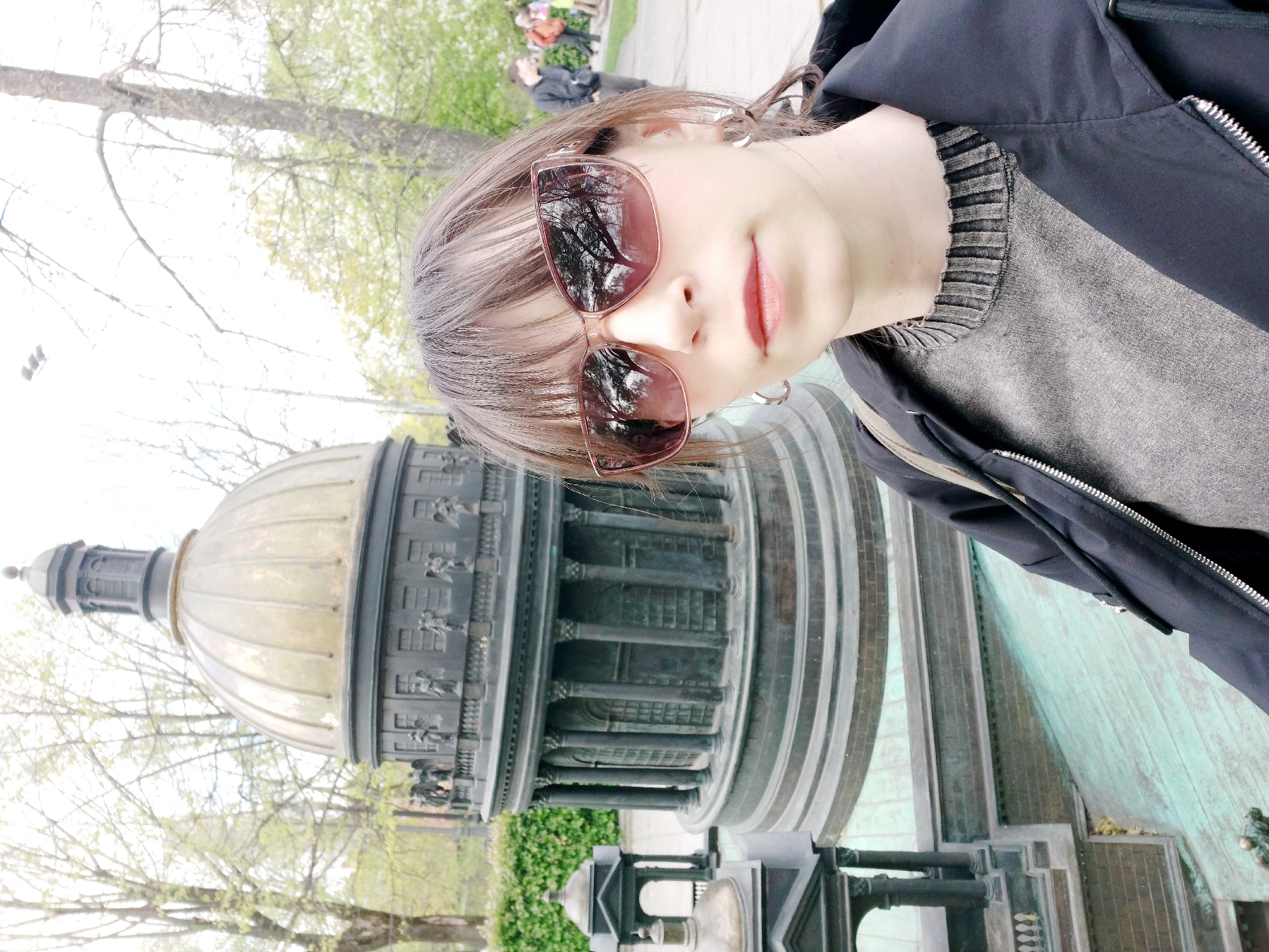 A woman wearing sunglasses, a gray top, and a black jacket takes a selfie in front of a miniature replica of St. Isaac's Cathedral in St. Petersburg, Russia. The dome of the miniature cathedral is visible behind her. Trees are visible in the background. People are also present in the far background.
