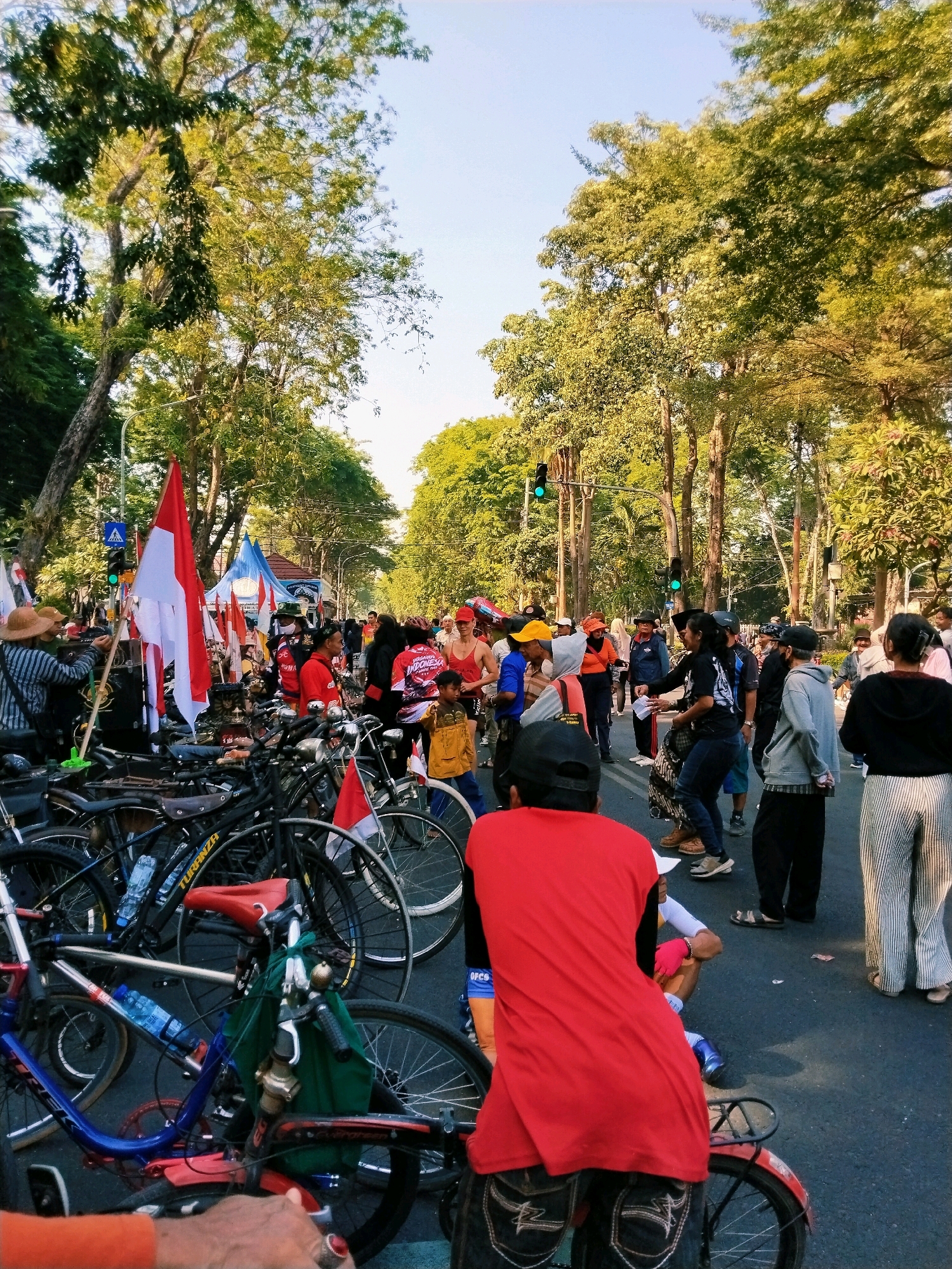 The image captures an outdoor scene in Surabaya, Indonesia, likely a street or a public gathering area, with dense foliage and trees lining the sides of the road. A crowd of people is gathered, many standing and some seated, suggesting an event or protest. Several bicycles are visible, parked or in the process of being moved, indicating a cycling-related activity or a mode of transportation for attendees.
Prominently displayed are Indonesian flags, red and white, along with other smaller flags, signifying a patriotic or nationalistic gathering. People are dressed in casual attire, with some wearing hats and masks. A few individuals are interacting, with some appearing to hand out or receive items. The atmosphere seems lively, with a mix of organized activity and spontaneous interaction. Traffic lights are visible in the background, suggesting the event is taking place on a road. The lighting suggests it is daytime, with sunlight filtering through the trees.