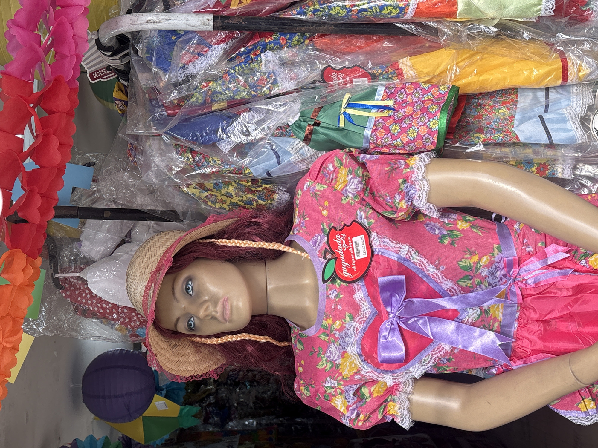 The image shows a mannequin wearing a traditional Brazilian "Festa Junina" costume, likely displayed in a shop or market stall in São Paulo, Brazil. The costume is a vibrant pink dress with a floral pattern, featuring lace trim and a large purple bow on the chest and at the waist. The mannequin also wears a straw hat with pink lace trim and has long, braided red hair.
Behind the mannequin, the scene is cluttered with more colorful clothing, many of which appear to be wrapped in plastic. These garments also display bright floral patterns, characteristic of Festa Junina attire. Festive decorations, such as colorful paper lanterns and garlands of red and orange paper flowers, hang above the display. The overall impression is one of a lively and colorful celebration, typical of the June Festivals held across Brazil. The specific location within São Paulo is not identifiable from the image, but the context suggests a commercial setting where these festive outfits are sold.