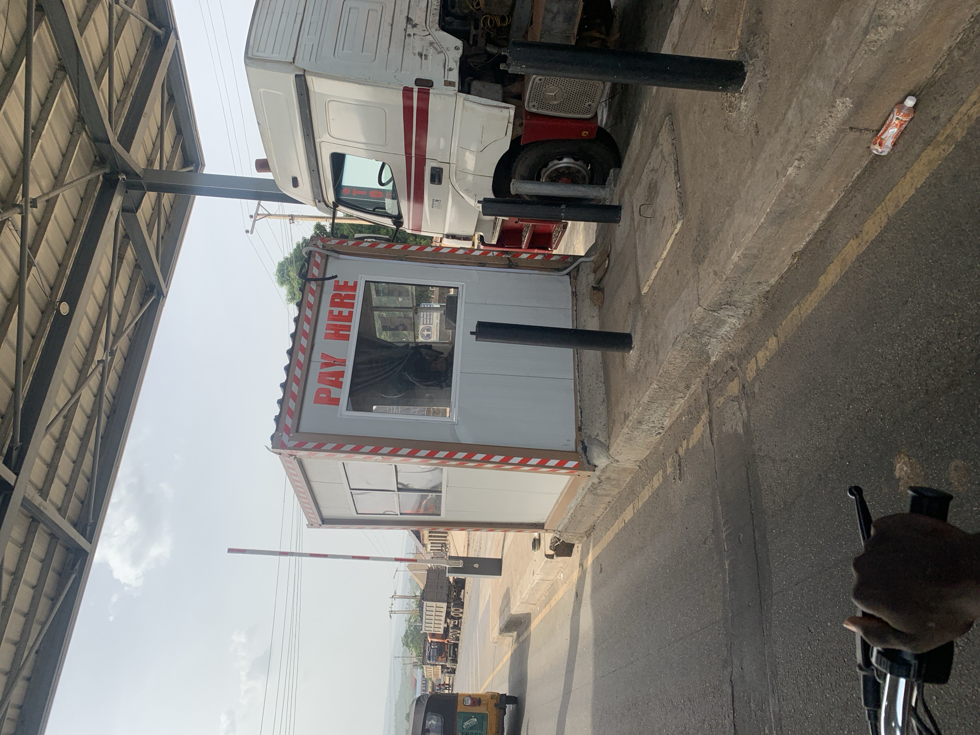 The image depicts a toll booth, labeled "PAY HERE" in red letters, situated on a road in Lokoja, Nigeria. A white and red truck is positioned beside the booth, partially obscuring the view of the structure. The toll booth is made of white panels with red and white striped trim. A barrier arm is in the down position in front of the booth. A bottle of Fanta Orange lies on the ground near the truck. A metallic canopy structure is seen above, partially covering the scene. In the background, there are more trucks and an auto-rickshaw. The image is taken from the perspective of someone on a moving motorcycle, as evidenced by a hand gripping the handlebar in the lower right corner. The weather appears to be hazy.