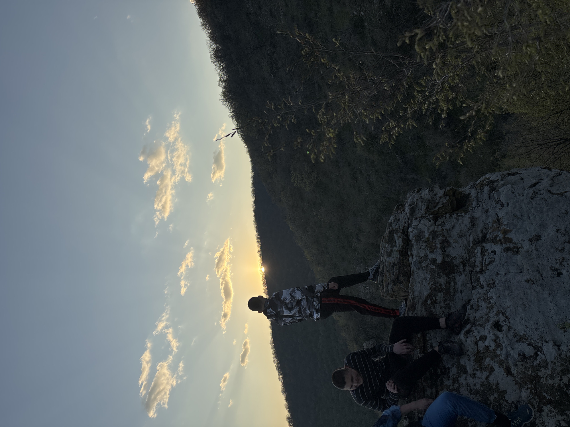 Three people are on a rock formation overlooking a valley. The sky is light blue with scattered clouds near the horizon, where the sun is setting. A dark mountain range is visible in the background. The location is near Goritsa, Bulgaria.