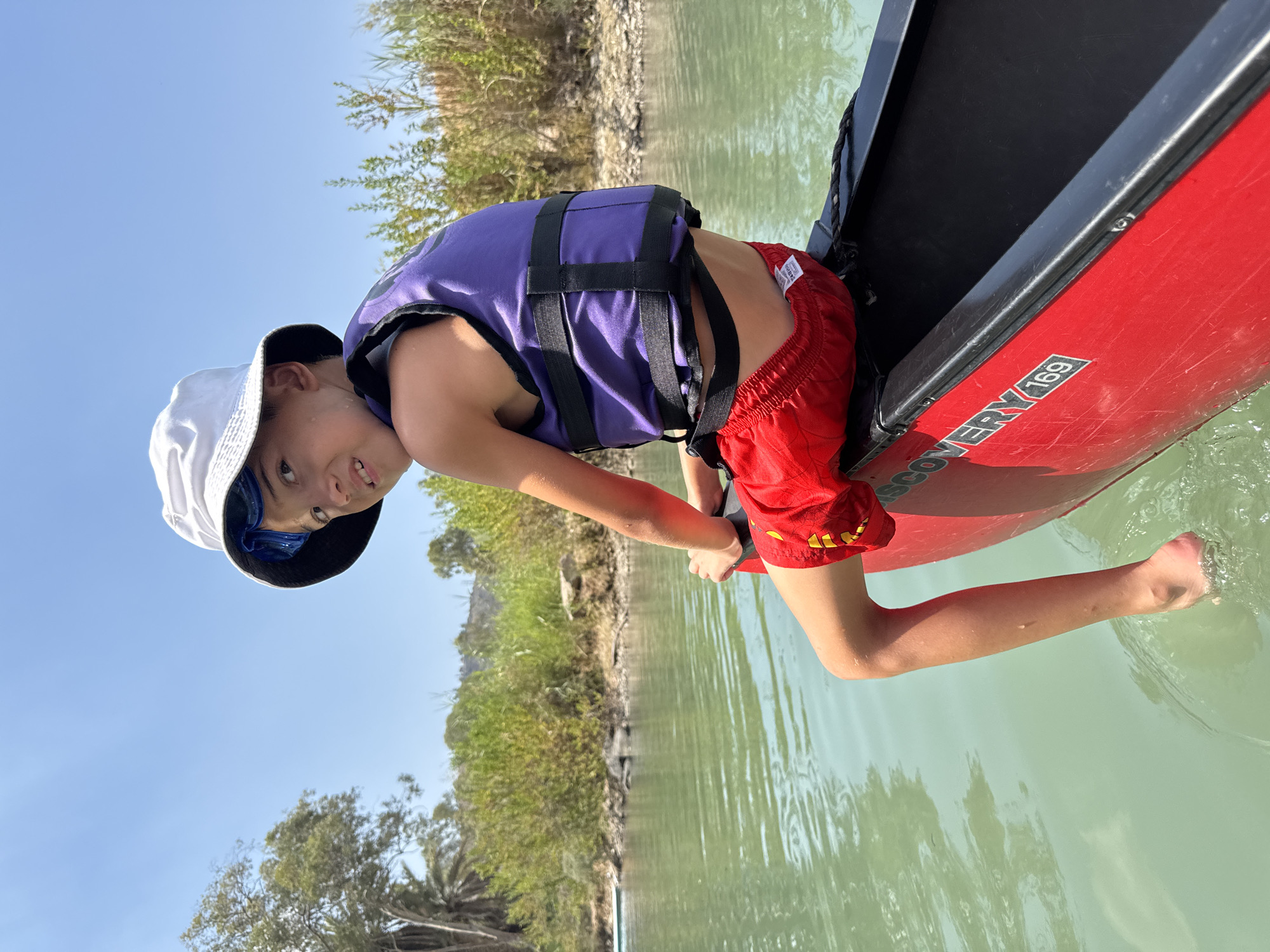 A young boy is leaning out of a red canoe with a black interior, his body positioned towards the camera. He wears a purple life vest over red swim shorts and a white bucket hat. Blue swimming goggles are perched on his forehead, visible under the brim of his hat. His face is turned towards the camera with a playful expression. His left arm is extended, with his hand submerged in the greenish water, while his right arm is bent and resting on the side of the canoe. The canoe appears to be on a body of water, likely a river or lake, with a rocky shoreline and lush green vegetation in the background. The sky is clear and blue. The scene suggests a sunny day, likely late morning or early afternoon, given the bright light and clear sky. A label on the side of the canoe reads "DISCOVERY 169". The setting is the Emek HaYarden Regional Council in Israel.