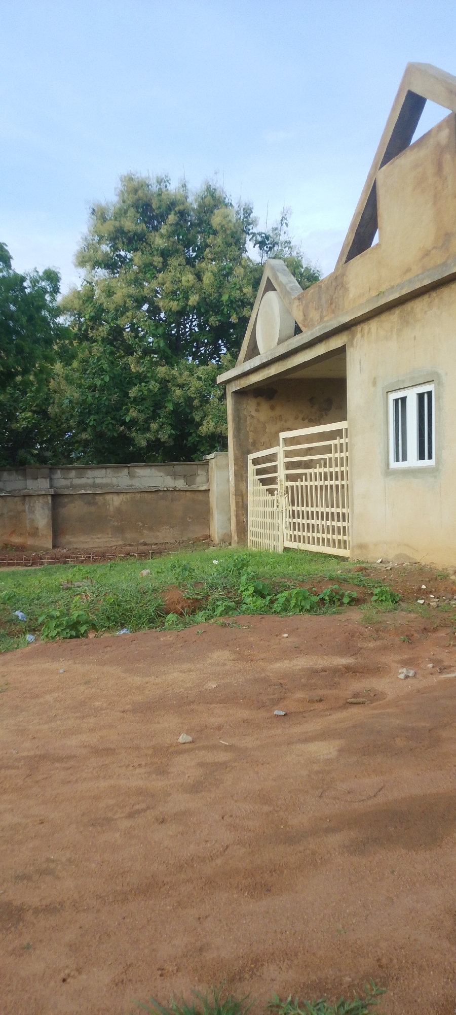 The image shows an unfinished building with a beige-colored exterior, featuring a distinctive triangular architectural element at the roofline and a circular decorative feature above an entrance. A white, barred gate is visible in front of the entrance, and a white-framed window is present on the building's wall. In the background, there is a concrete block wall, and beyond that, lush green trees, one of which is laden with light-colored flowers. The ground in the foreground is a reddish-brown dirt path, with patches of green grass and small stones scattered about. The sky is a pale blue, suggesting it is daytime, possibly late afternoon or early morning given the soft light. The scene appears to be in a semi-rural or developing area, consistent with a location like Jalingo, Nigeria, where construction and natural elements coexist. There are no people visible, and the overall impression is one of ongoing construction and a natural, perhaps slightly unkempt, environment.
