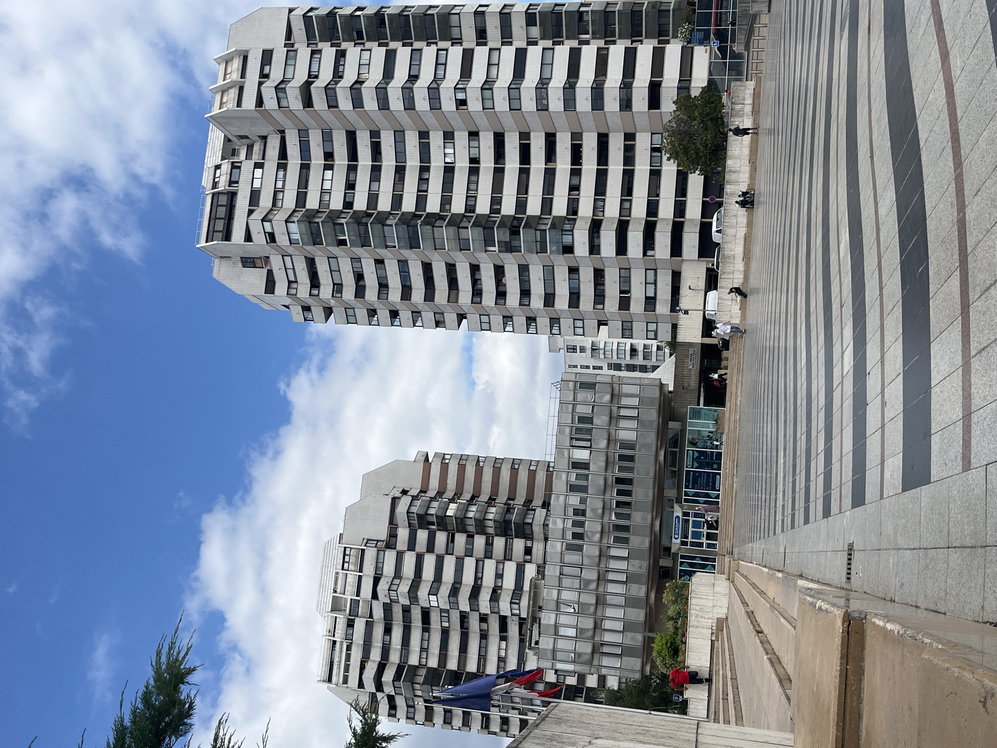 The image shows a view of the Place de l'Échutier in Créteil, France, with two tall, modern buildings dominating the skyline. The building on the right is a multi-story residential tower with a distinctive facade featuring numerous balconies and windows. The building on the left, though shorter, also exhibits a similar architectural style with multiple levels and windows.
In the foreground, a wide plaza with a distinctive black and white striped pavement pattern extends towards the buildings. To the left of the plaza, there are concrete steps and a ramp leading up to the entrance of one of the buildings. Two flags, appearing to be the French flag and another national flag, are displayed on flagpoles near the entrance.
Several people are visible in the scene. On the steps to the left, a person in a red shirt is ascending. In the plaza, individuals are seen walking, sitting, and interacting. Some are gathered near the entrance of the building, while others are further out in the plaza. A person on a scooter is visible in the middle ground. The sky above is blue with scattered white clouds. The overall environment suggests an urban public space with residential and possibly commercial buildings.