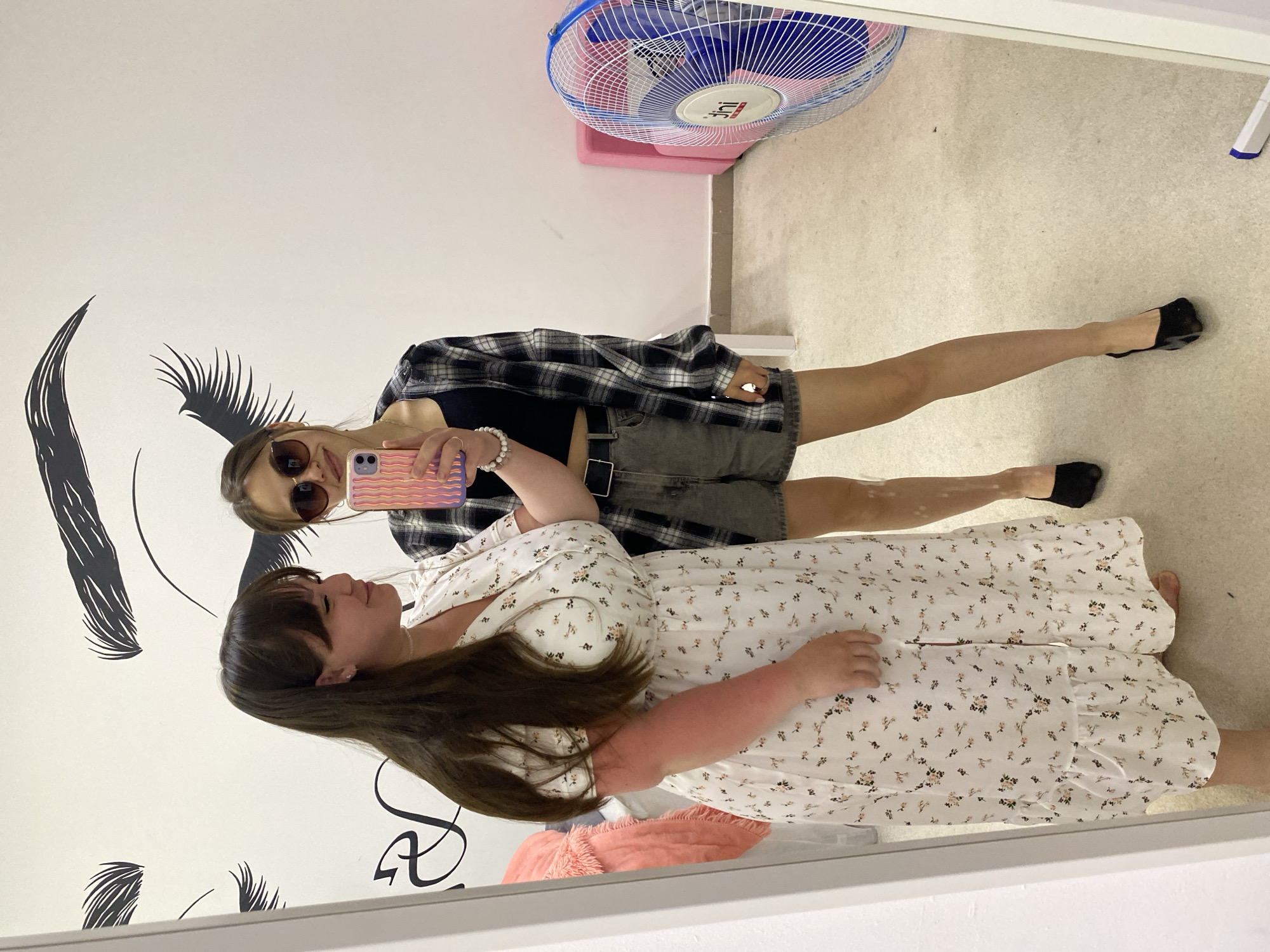Two young women are posing for a mirror selfie in what appears to be an interior setting. The woman closer to the mirror is wearing a white dress with a floral pattern. The other woman is wearing a black top, a plaid shirt, grey shorts, and sunglasses. She is holding a phone and taking the picture. In the background, there is a white wall with an illustration of an eye and eyelashes. A fan is visible in the upper right corner of the image. The exact location in Votkinsk, Russia is not possible to determine from this image.