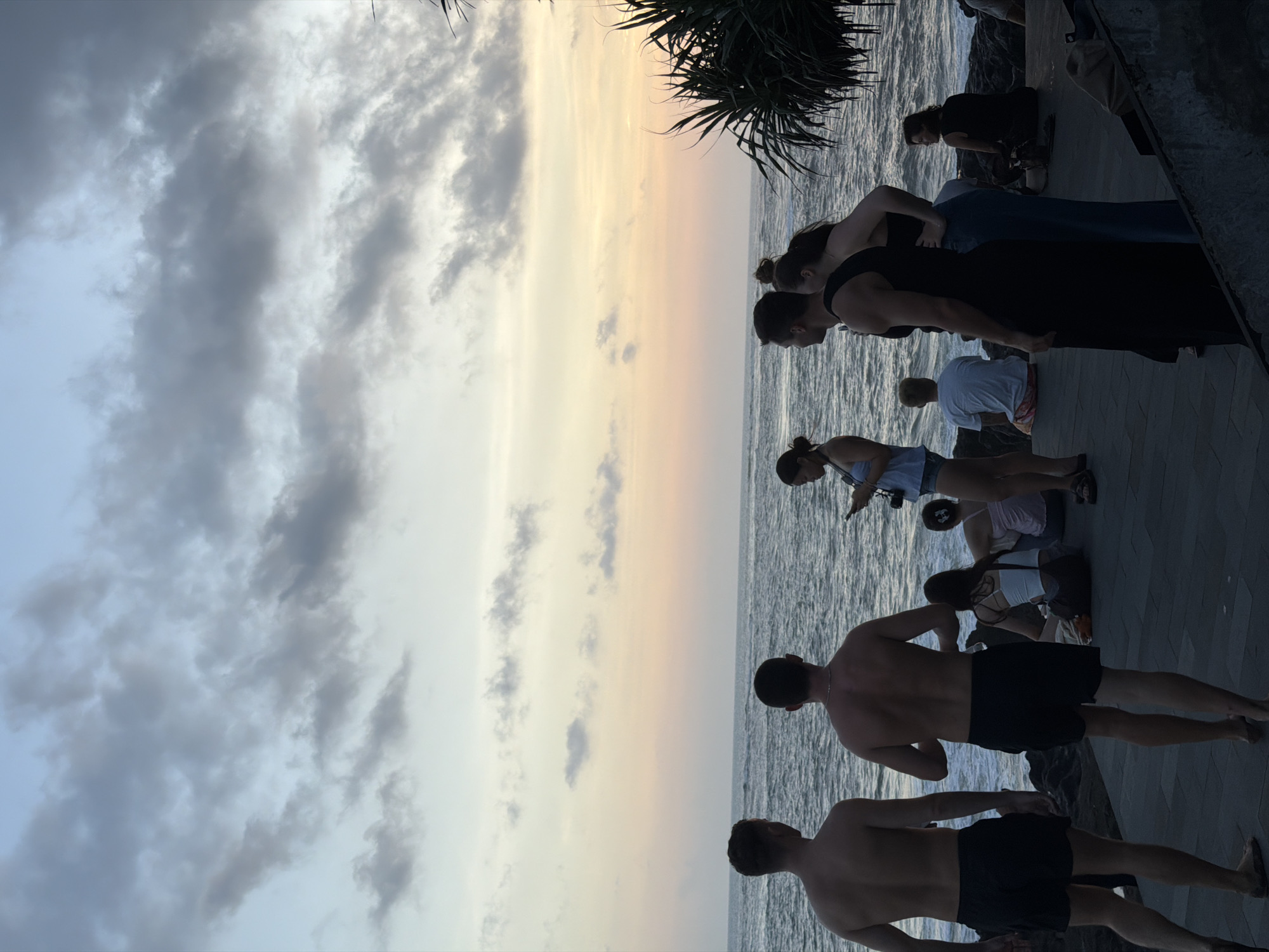 The image shows a group of people at Tanah Lot temple during sunset in Mengwi, Indonesia. Two shirtless men in black shorts stand near the edge of a paved pathway. Behind them, a group of people of various ages and attire stand, some looking toward the ocean. To the right of the pathway is a sparse bush. Above the ocean is a light-colored sky with clouds.