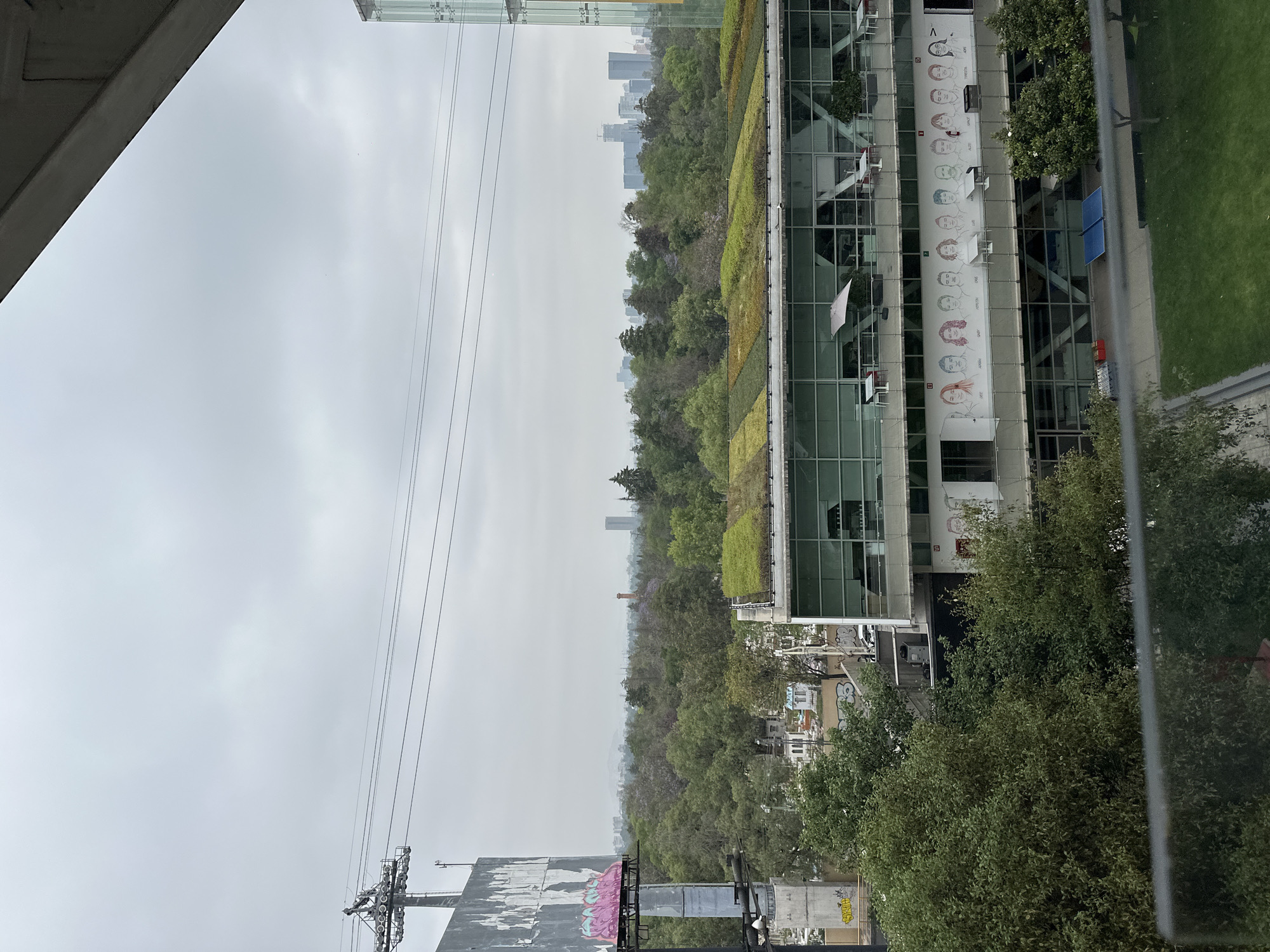 This is an elevated view of a modern urban landscape in Mexico City on an overcast day.
In the upper left corner, a large concrete structure, possibly part of a bridge or overhead walkway, partially frames the scene. Thin utility lines, likely power or tram wires, stretch horizontally across the upper portion of the grey, uniformly cloudy sky.
The midground features prominent architectural elements and green spaces. A long, modern building with a distinct green roof (a living roof or garden) runs horizontally across the image. Adjacent to it, to the right, is another contemporary building characterized by large glass windows. This building displays a notable mural or banner on its side, featuring a horizontal series of stylized cartoon faces, each with a name printed above it, such as "MIGUEL," "ARTURO," "DAVID," "SARA," and "URBANO." A white flag hangs from this building. Below these structures and to the right, a paved path runs alongside a manicured green lawn, where two blue rectangular objects (possibly solar panels) are visible.
A dense area of trees and vegetation, resembling a park or a large urban green space, separates the foreground buildings from the background. Beyond this treeline, a hazy distant cityscape with numerous tall buildings is visible, suggesting a sprawling urban environment.
In the lower left, a large, weathered billboard or sign structure is partially visible, adorned with pink graffiti and some indistinguishable text, with what appears to be "SERGIO" and possibly "URBANO" scrawled on it.
The overall weather is cloudy and overcast, indicating daytime without direct sunlight. No people or specific activities are clearly visible, presenting a static snapshot of the city.