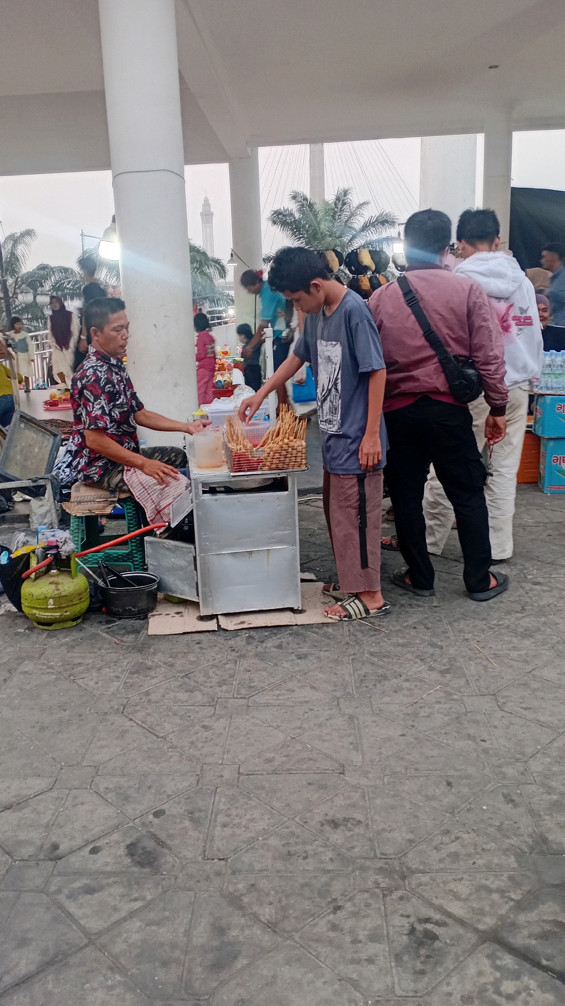 The image captures a street food vendor operating a small mobile cart outdoors in Jambi City, Indonesia. The vendor, a man with short black hair, is seated on a stool behind his cart, wearing a dark patterned shirt. He appears to be serving a customer, a young man wearing a grey t-shirt and brown pants, who is reaching for food from the cart.
The food cart is made of metal and contains a basket of skewered food items, likely satay or meatballs, and a clear container with sauce. To the left of the cart, a green LPG gas cylinder and a black bucket are visible. In the background, other people are present, suggesting a public area, possibly a park or market. The ground is paved with square tiles. Beyond the immediate scene, a white pillar and a suspension bridge structure are visible, with palm trees scattered in the environment. The lighting suggests it is daytime, possibly overcast.