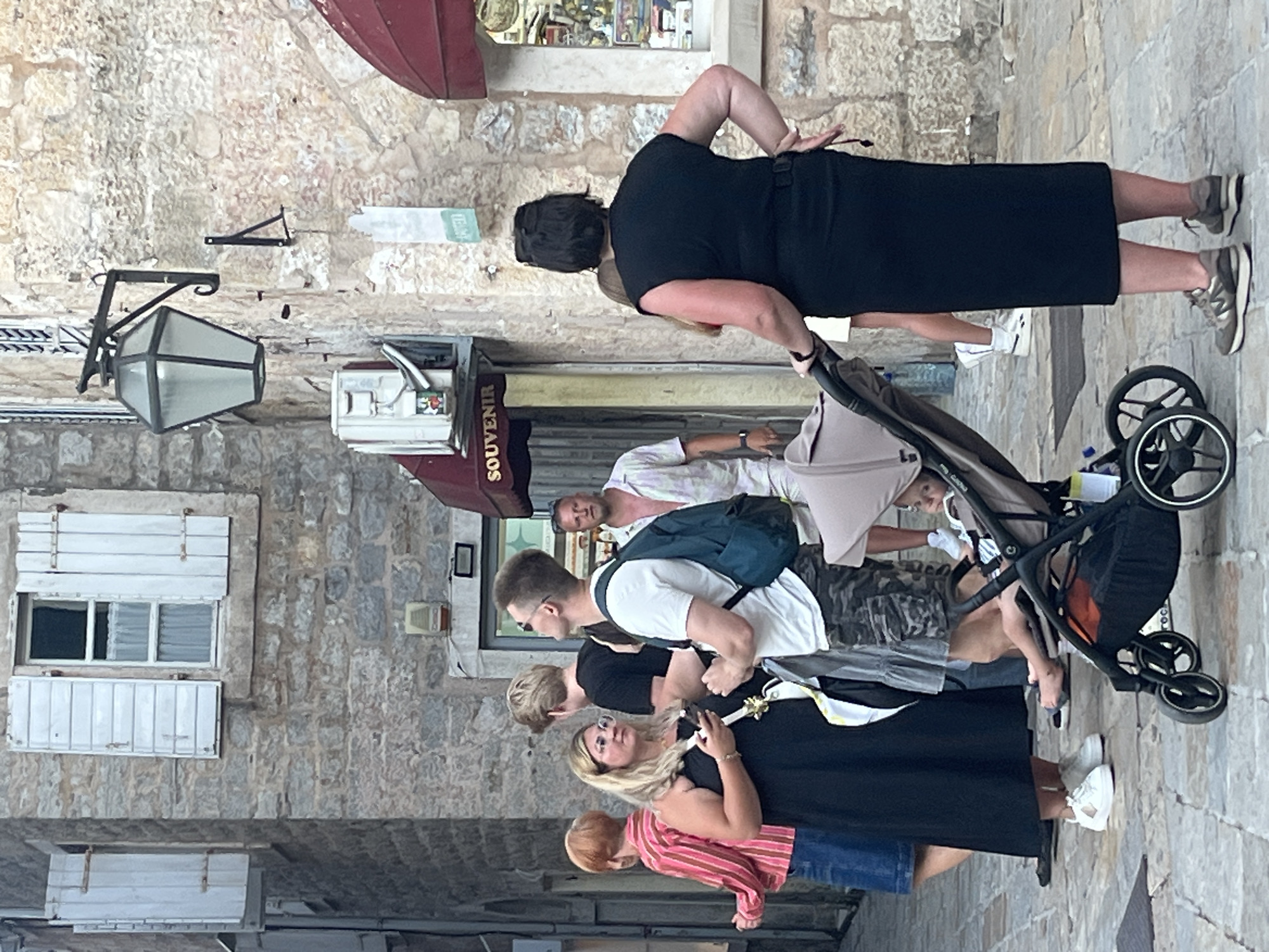 The image depicts a street scene in Budva, Montenegro, likely within the old town, characterized by its narrow cobblestone street and traditional stone architecture. In the foreground, a woman in a black dress pushes a stroller with a baby inside. To her left, another woman in a black dress, wearing glasses, holds a smartphone. Behind them, a man with a backpack stands looking upwards, and another man in a striped shirt is partially visible. Further back, a man in a light-colored patterned shirt and shorts stands looking towards the group. The background shows a building with stone walls and windows with white shutters, an outdoor lamp fixture, and a shop with a red awning and a "SOUVENIR" sign. The overall atmosphere suggests a casual, daytime outing in a historic urban environment.