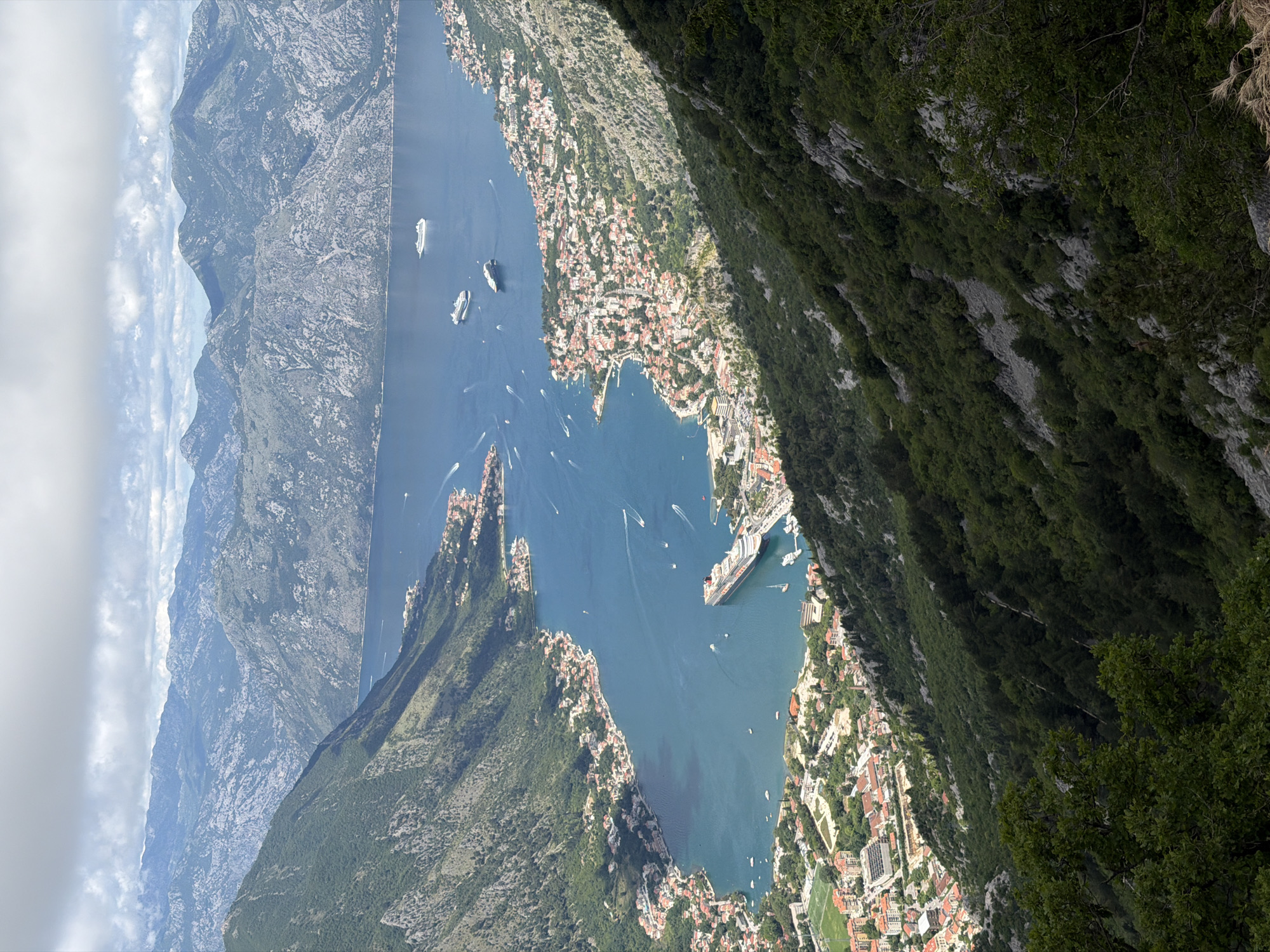 A wide-angle, high-altitude panoramic view captures the stunning Bay of Kotor in Montenegro, framed by steep, rugged mountains under a partly cloudy sky. The deep blue, fjord-like bay winds through the center of the image, from the foreground towards the distant background.
Along the shores of the bay, numerous picturesque towns and settlements are nestled, their buildings characterized by red and orange tiled roofs, extending from the water's edge up the lower slopes of the mountains. A large, multi-deck cruise ship with a dark hull and white superstructure is prominently anchored or docked in a town on the right side of the bay. Several other large white yachts or smaller cruise ships are visible further out on the water, alongside numerous smaller white boats, some leaving noticeable white wakes indicating movement.
The mountains enveloping the bay are heavily forested with lush green trees and shrubs, particularly in the foreground and on the closer slopes. In contrast, the more distant mountain ranges in the background appear grayer and rockier, with patches of lighter vegetation. The sky above is a mix of bright blue and scattered white clouds. The scene is illuminated by bright daylight, suggesting a clear day, likely late morning or early afternoon. The location, Špiljari, Montenegro, offers this breathtaking vista of the iconic Bay of Kotor. No people or text are discernible in this expansive scene.