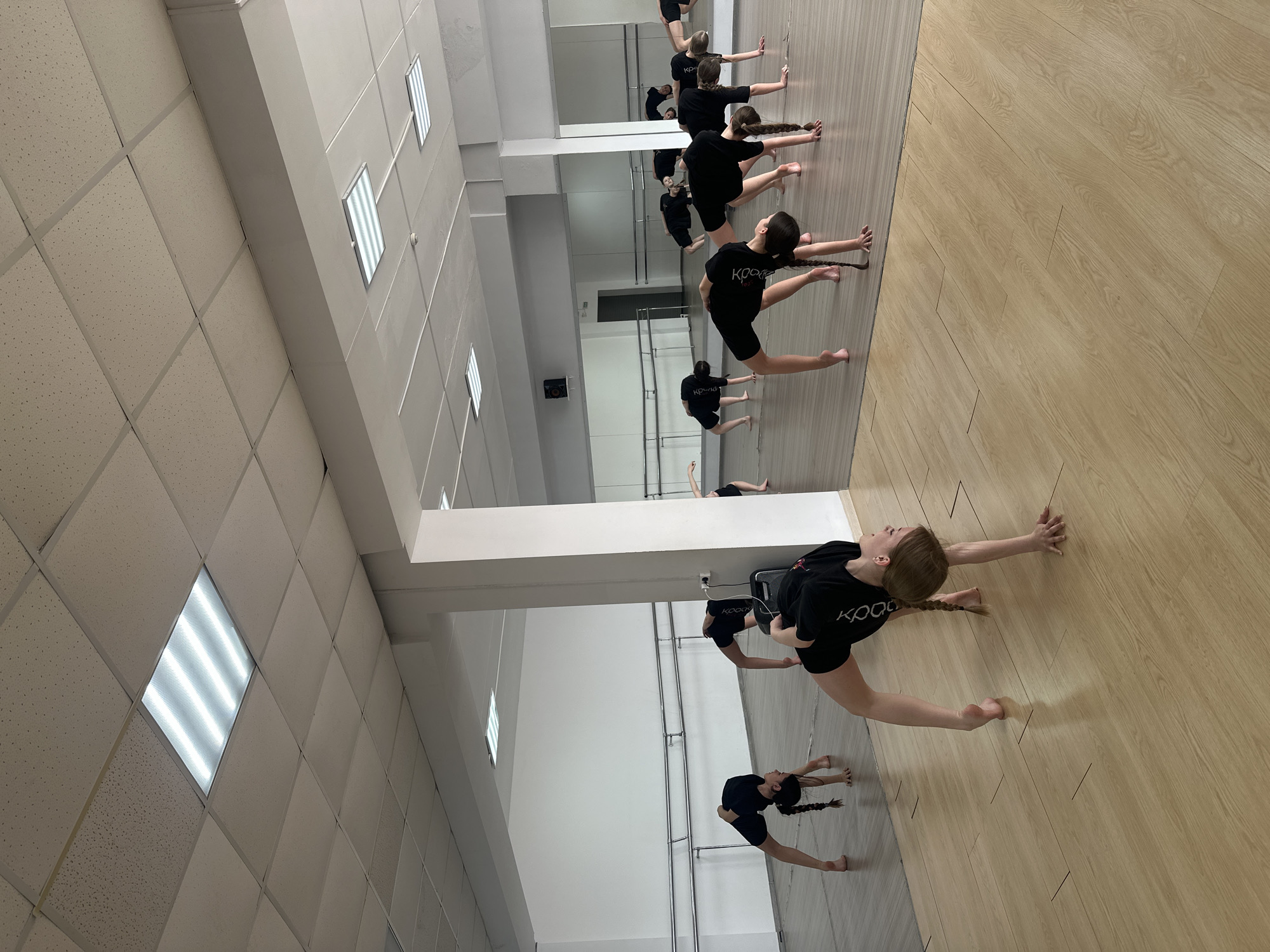 A horizontally oriented photograph shows several girls in a dance studio in Votkinsk, Russia. The dancers are wearing black shirts and shorts and are practicing a stretch, leaning forward with arms extended, legs in a split position. A mirror runs along the wall, reflecting the dancers. The studio has a wooden floor and a white tiled ceiling with fluorescent lights.