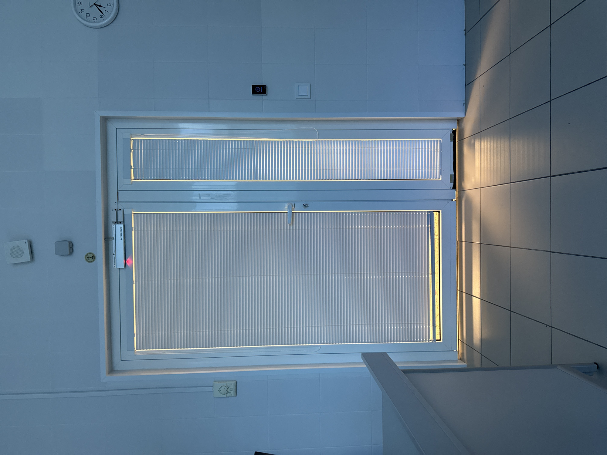 The image shows a room with white tiled walls and floor. There is a double window in the center, covered with horizontal blinds. The window is white framed. Light is streaming in from the right side of the image, casting a shadow on the tiled wall and floor. Various objects are visible on the wall to the left of the window, including a speaker, a sensor, and a small control panel. There is a clock in the upper left corner and a light switch and electrical outlet above the window. A counter or shelf is partially visible in the lower right corner. The image does not provide enough information to identify the location as Moscow, Russia.