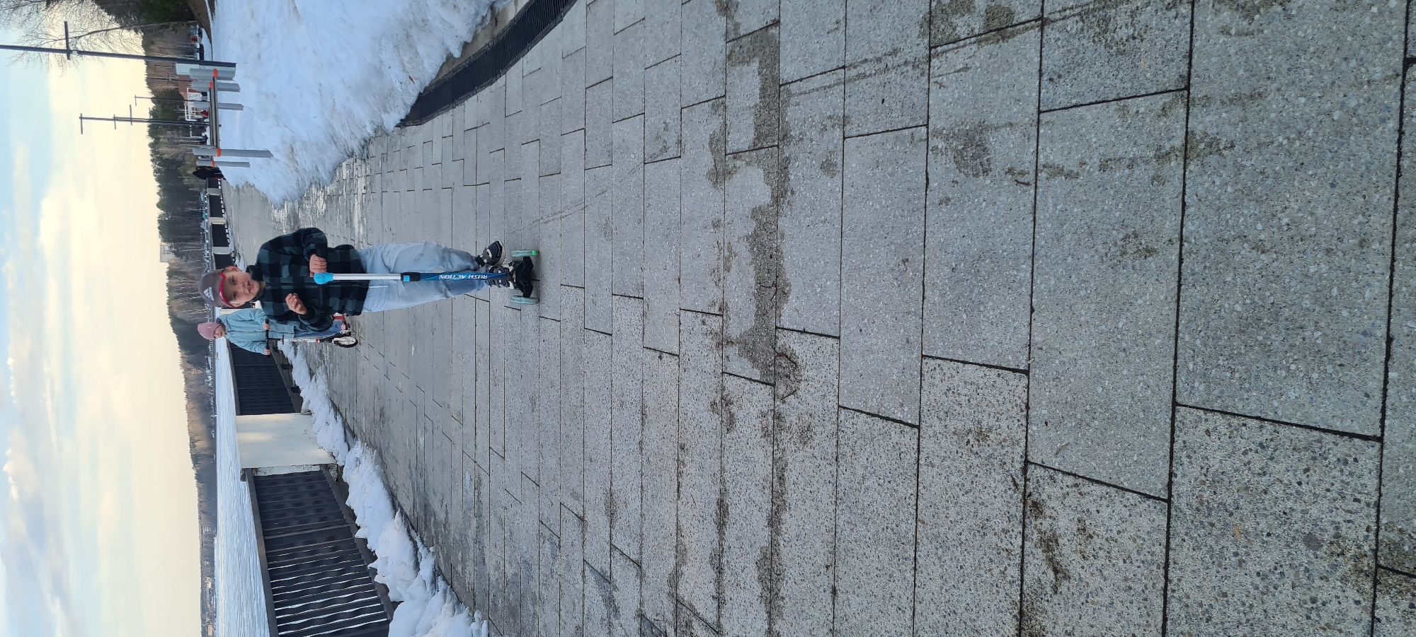 A photograph captures a scene along a paved walkway in Chaikovsky, Russia. A boy is standing on a scooter. Another person is behind him. Snow remnants are visible along the edge of the walkway, adjacent to a body of water. Railings separate the walkway from the water. In the distance, poles and bare trees can be seen. The sky is pale. The walkway is composed of rectangular tiles.