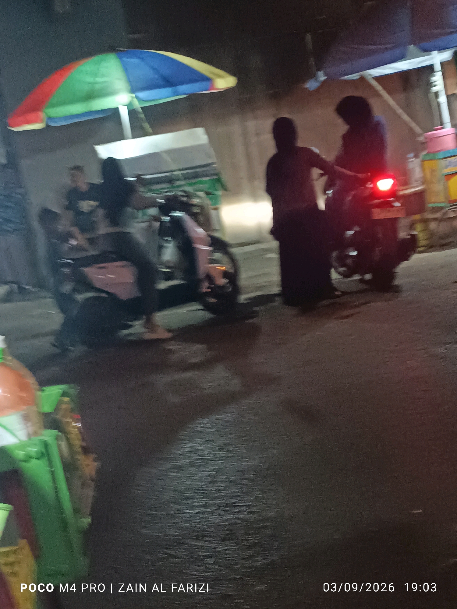 In Buduran, Indonesia, a scene unfolds at night featuring a colorful umbrella providing shade over a street or market area. Two individuals are present, one seated on a scooter and another standing nearby. The scooter has its taillight illuminated, casting a red glow. To the left, a green cart or display is partially visible, holding what appear to be bottled drinks. The ground is asphalt, and the overall lighting suggests an urban environment at dusk or night.