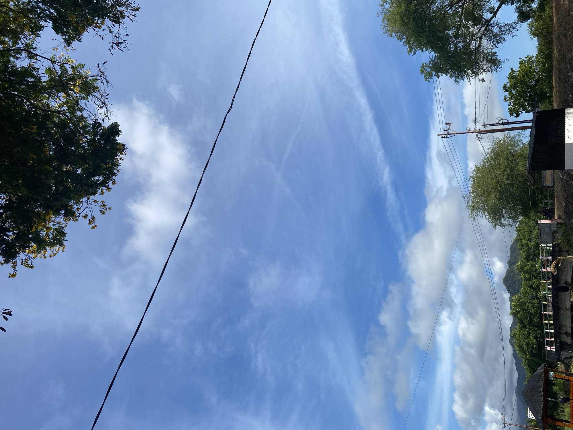 In Aneuk Paya, Indonesia, a clear blue sky with scattered white clouds dominates the upper portion of the image. A thin black power line bisects the sky diagonally from the upper left to the lower right. In the lower half of the image, a bridge with colorful railings spans a water feature. To the left of the bridge, a thatched-roof shelter with wooden supports stands near the water. Dense green trees and vegetation line the background, with rolling hills or mountains visible in the distance. Power poles and additional power lines are also present, indicating a developed area within the natural landscape. The overall scene depicts a sunny day in a semi-rural or coastal setting.