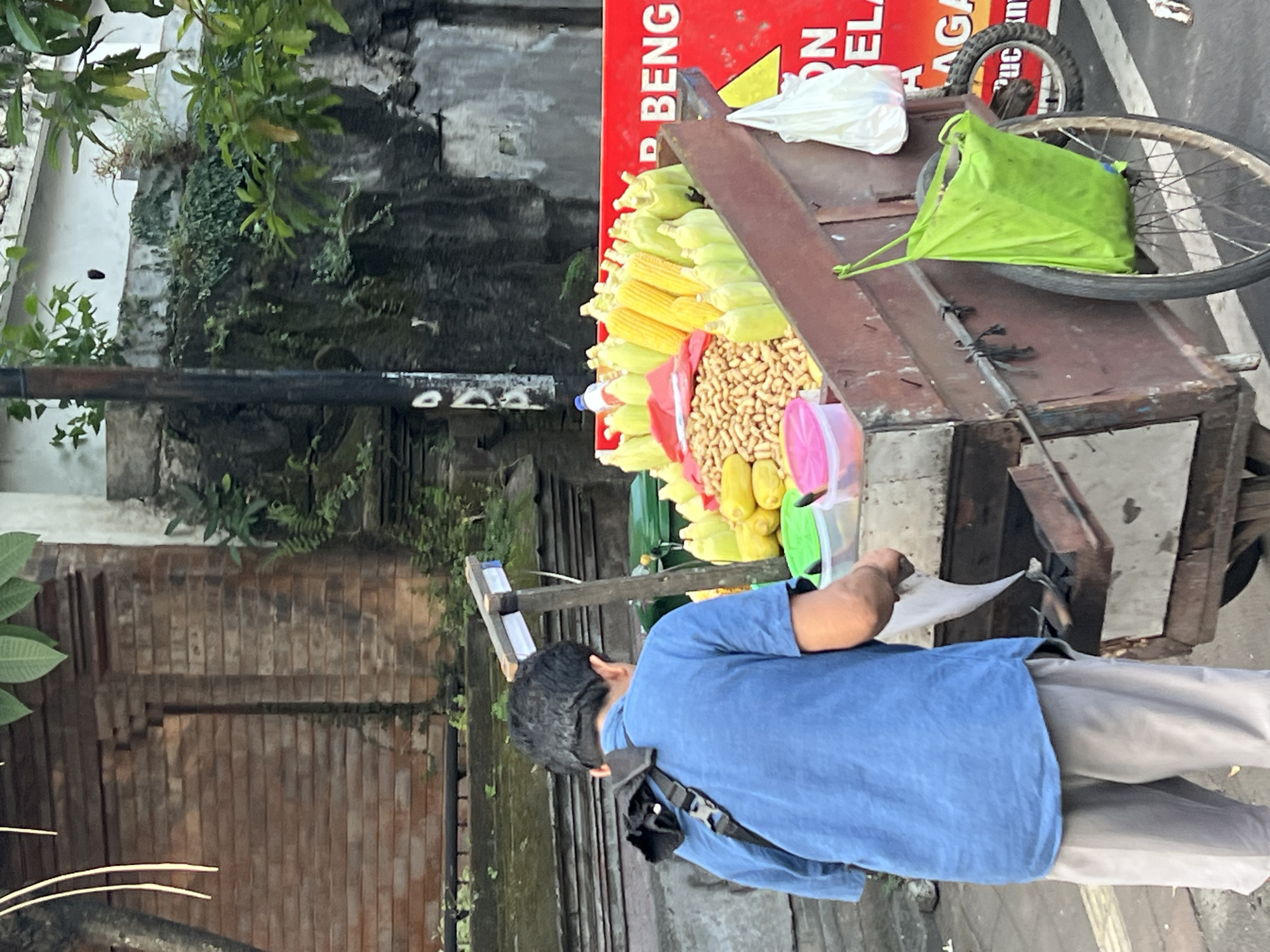 A man in a blue t-shirt and light-colored shorts stands with his back to the camera, facing a food cart filled with corn and peanuts. The cart is made of wood and metal, with two large spoked wheels. A bright red sign with white lettering is attached to the back of the cart. A green bag is hanging from the side of the cart. The background shows a brick wall with greenery growing on it and a pole. The scene appears to be on a street in Denpasar, Indonesia, likely a market or roadside vendor area.