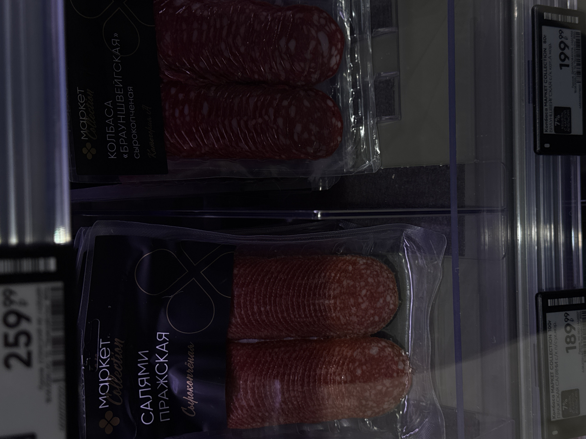 The image shows a close-up view of two packages of sliced sausage products in a supermarket display case. The package on the left contains "САЛЯМИ ПРАЖСКАЯ" (Prague Salami), and the package on the right contains "КОЛБАСА «БРАУНШВЕЙГСКАЯ»" (Braunschweig Sausage). Both products are from the "Market Collection" brand. The sausages are visible through the clear plastic packaging, with the Prague Salami appearing to be slightly more reddish and less marbled than the Braunschweig Sausage. Below the product images, price tags are visible. The price tag for the Prague Salami shows 189.99 rubles, and the price tag for the Braunschweig Sausage shows 199.99 rubles. The background is dark, and some reflections from the display lighting are visible. There are no people or animals present in the image. The scene appears to be a retail environment, likely a section of a grocery store in Moscow, Russia, focusing on processed meat products.