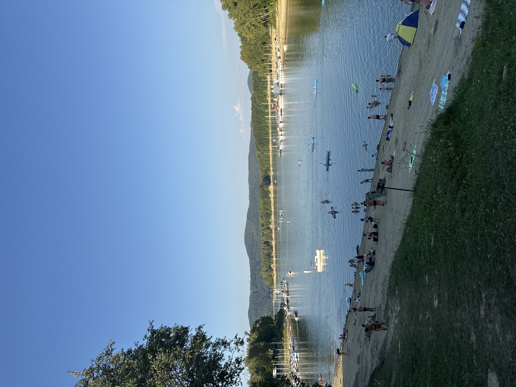 The image depicts a scenic view of the Solina Lake in Polańczyk, Poland, on a clear, sunny day. A sandy beach lines the foreground, with several people relaxing and sunbathing on towels and blankets. Some individuals are wading in the shallow water, while others are swimming further out. A green and yellow tent is pitched on the beach.
The lake itself is calm, with numerous sailboats and yachts docked in a marina to the left and further along the shore. Several individuals are engaged in water activities, including kayaking and paddleboarding. The water reflects the clear blue sky and the surrounding lush green forested hills.
In the background, a distant bridge can be seen spanning the lake, and the hilly landscape is dotted with trees. The overall atmosphere is one of a peaceful, recreational summer day.
