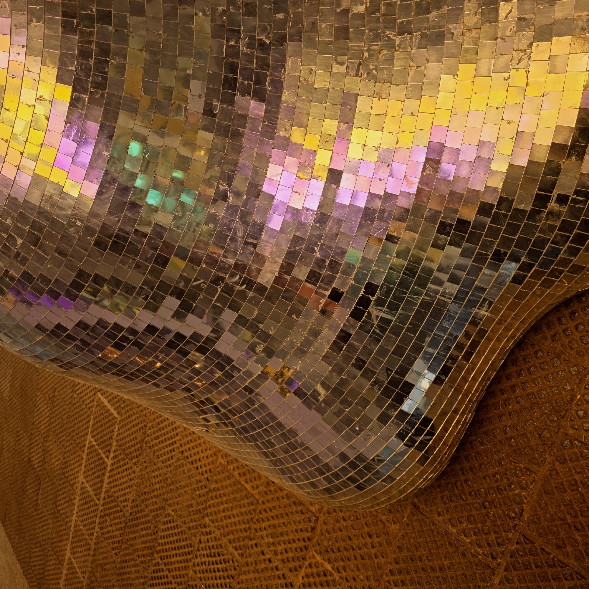 The image shows a section of the lobby of the Okhta Mall shopping center in Saint Petersburg, Russia. The curved ceiling is covered in small square mosaic tiles that reflect a spectrum of colors including gold, yellow, pink, and iridescent hues. Below the mosaic ceiling, there is a patterned surface, possibly another section of wall or the floor, that features a grid-like design in a brown or bronze color. The lighting in the scene appears to be warm, enhancing the golden tones of the mosaic and the lower surface.