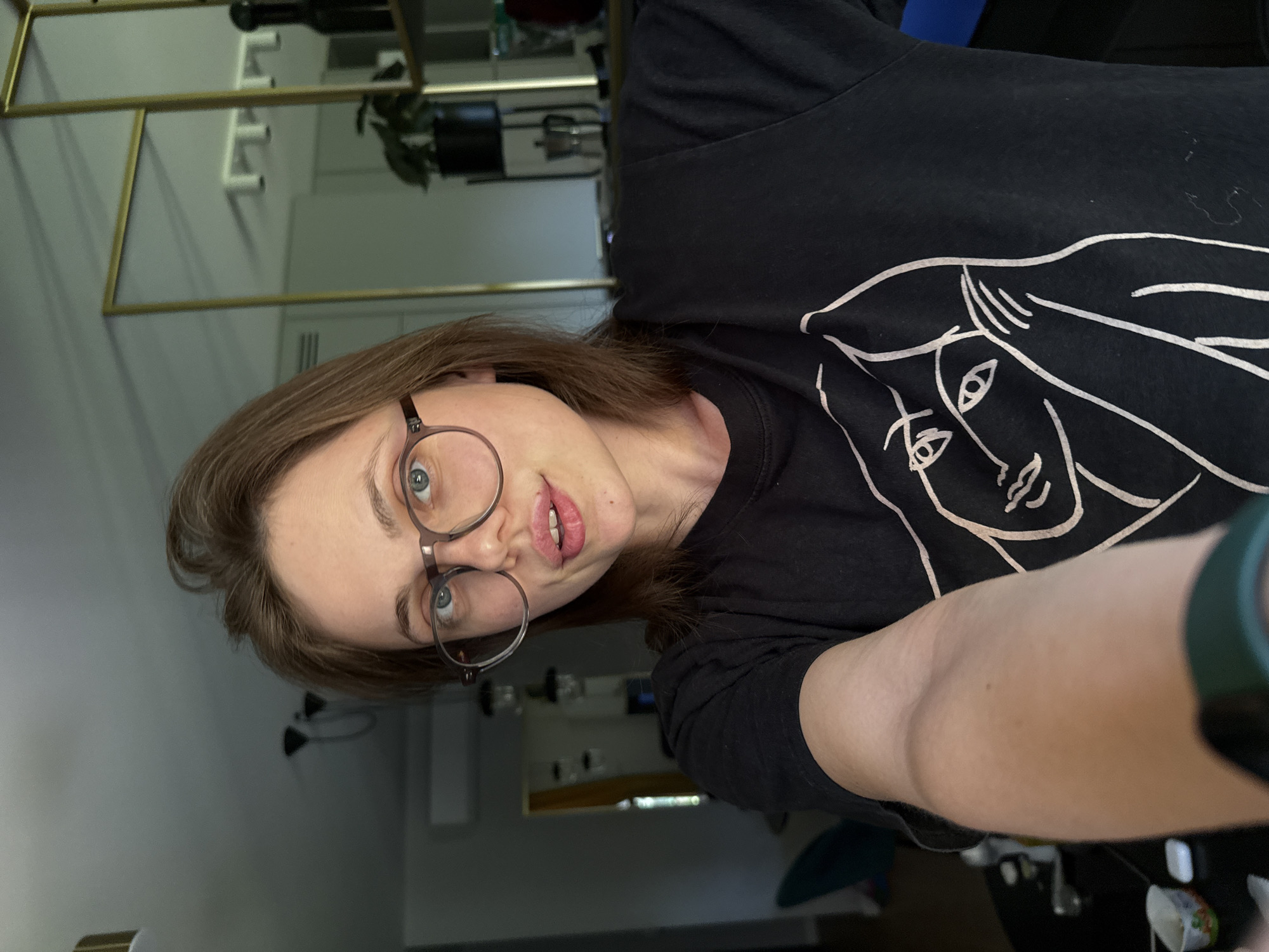 The image is a vertical, close-up selfie of a person with shoulder-length brown hair and light skin. They are wearing round, thin-rimmed glasses and a black t-shirt with a white graphic of a face. The person's mouth is slightly open, revealing their teeth and tongue, and they appear to be looking at the camera with a slight smirk. The background is blurry and shows a room with a mirror on the wall, reflecting shelving units and some plants. The lighting suggests it could be daytime. The person is wearing a dark green fitness tracker on their left wrist. The overall impression is casual and informal.