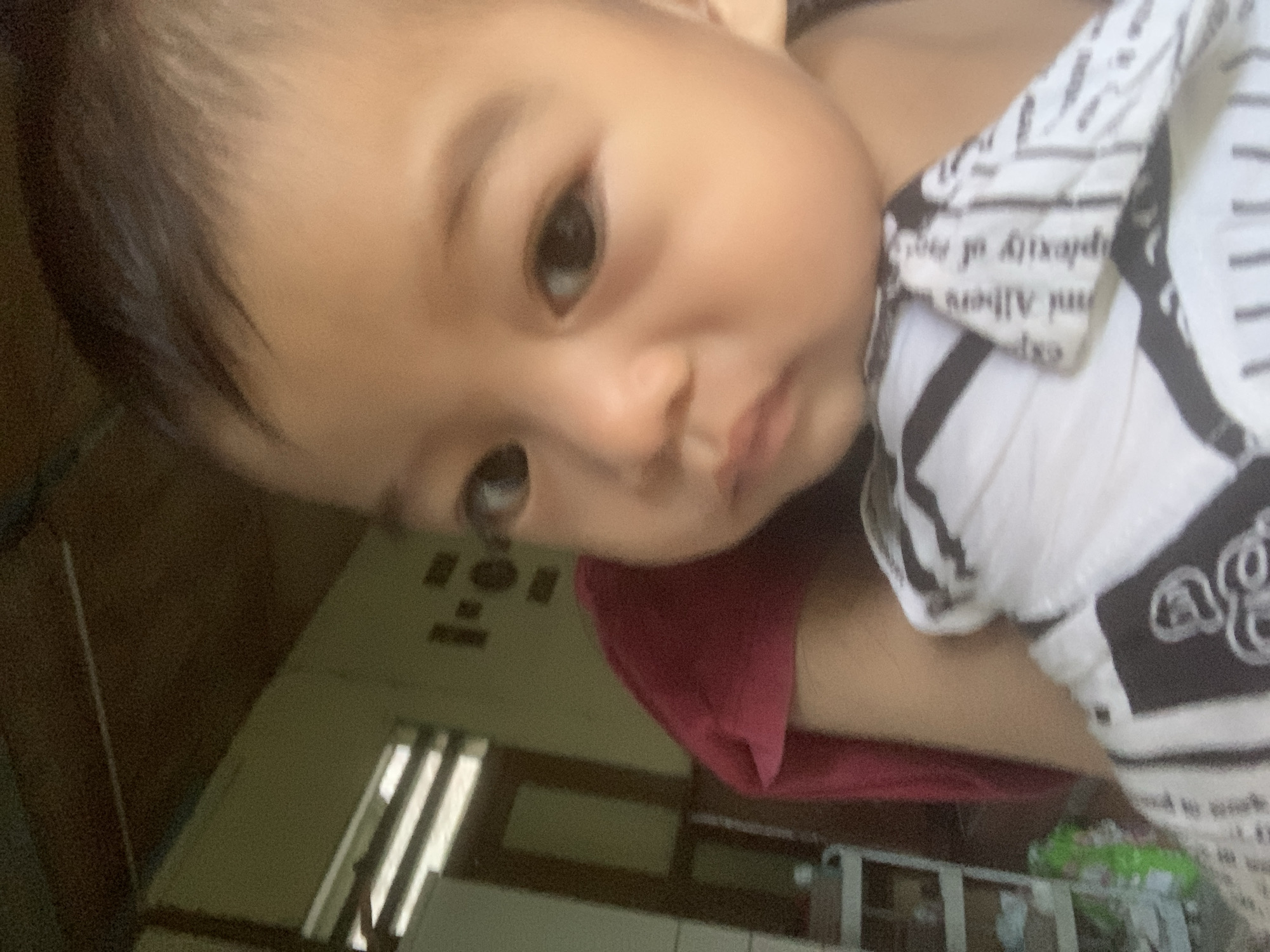 A close-up, slightly blurred photograph depicts a young child with dark hair and eyes. The child is wearing a white shirt with black stripes and text patterns. The child is held, with a red garment visible behind the child's neck. The background appears to be an interior space with a window showing daylight. The location is Sungai Meriam, Indonesia, but specific details about the location within the city are not discernable.