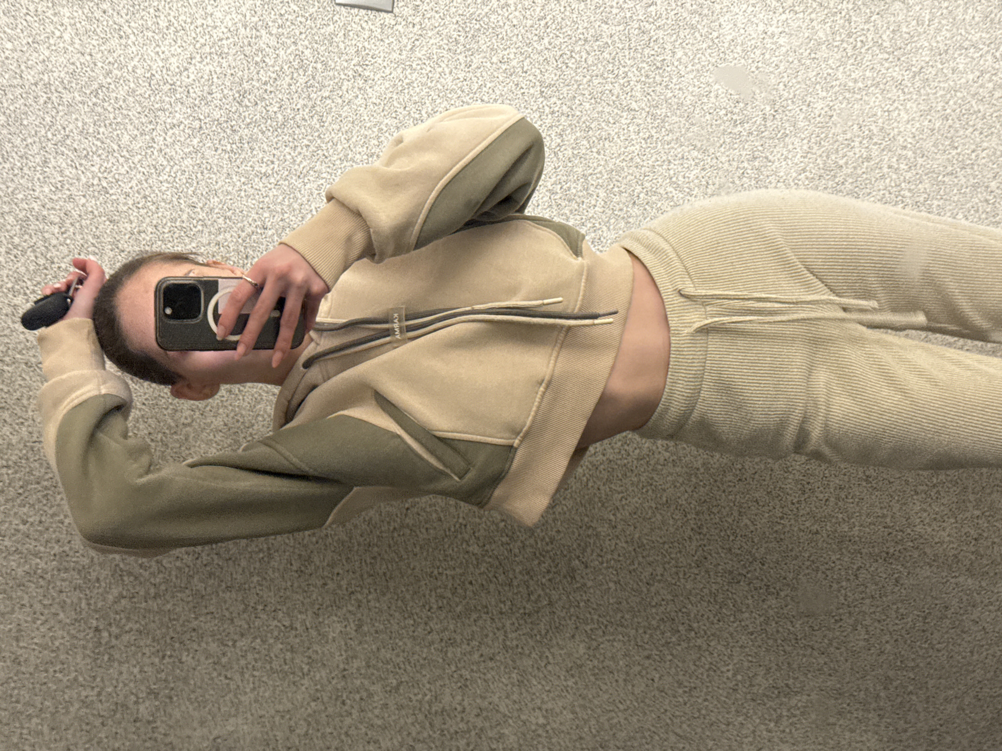 A person is seen in a selfie-style photograph indoors, presumably in an elevator with a gray speckled wall. The person is holding a black smartphone in their right hand to take the picture, while the left hand holds a car key. They are wearing a tan tracksuit with contrasting green panels on the sleeves and pockets of the jacket, which is a cropped zip-up style. The person's face is mostly obscured by the phone. The tracksuit pants appear to have a ribbed texture. The lighting is muted, and the focus is on the person and their attire. There is no clear indication of the location being in Sofia, Bulgaria, or any other city.
