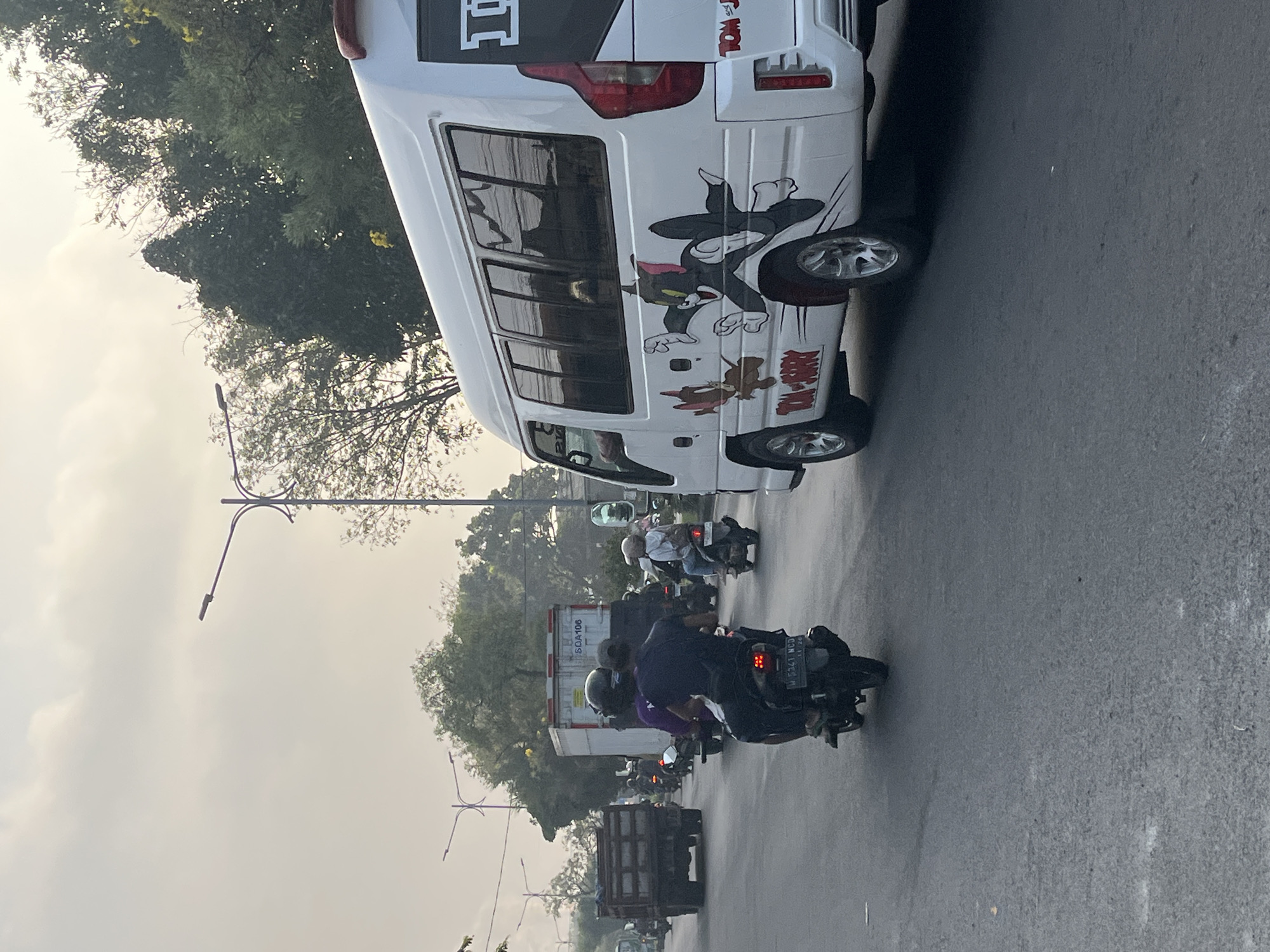 The image captures a street scene in Gelam, Indonesia, likely during the late afternoon due to the diffused sunlight and overcast sky. The foreground is dominated by a paved road filled with various vehicles. In the right foreground, a white minibus is prominently featured, adorned with a cartoon decal of Tom and Jerry. To the left of the minibus, a dark-colored motorcycle carries two individuals, the passenger in the back wearing a purple shirt and a helmet. Further down the road, more motorcycles are visible, along with a large, gray truck with a white container and markings that read "SQA106." A darker, older-model truck with a wooden bed is also present in the mid-ground. Streetlights are visible in the background, and dense foliage lines the side of the road, suggesting a city environment with significant greenery. The overall atmosphere appears to be that of moderate traffic congestion.